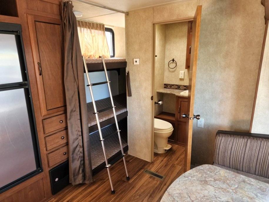 An RV rental interior features practical bunk beds, a ladder, a compact bathroom, and a partial kitchen with a fridge.. Forest River Wildwood 2014