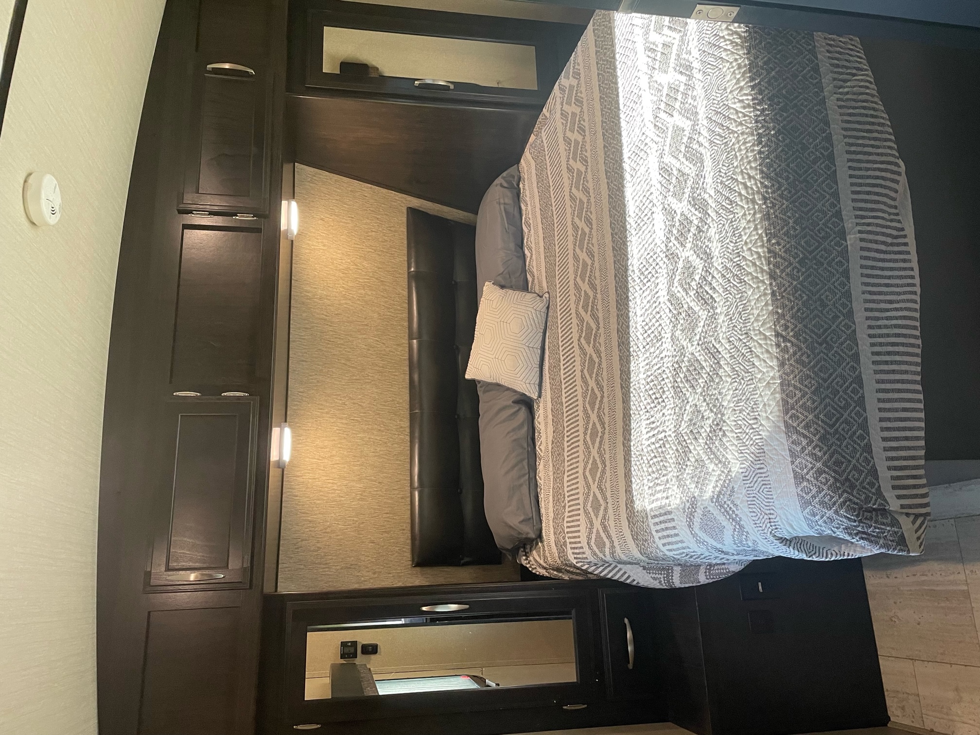 Inside an RV rental, a well-appointed bedroom with a comfy bed, patterned quilt, dark wood cabinets, and integrated storage.. Grand Design Imagine 2018