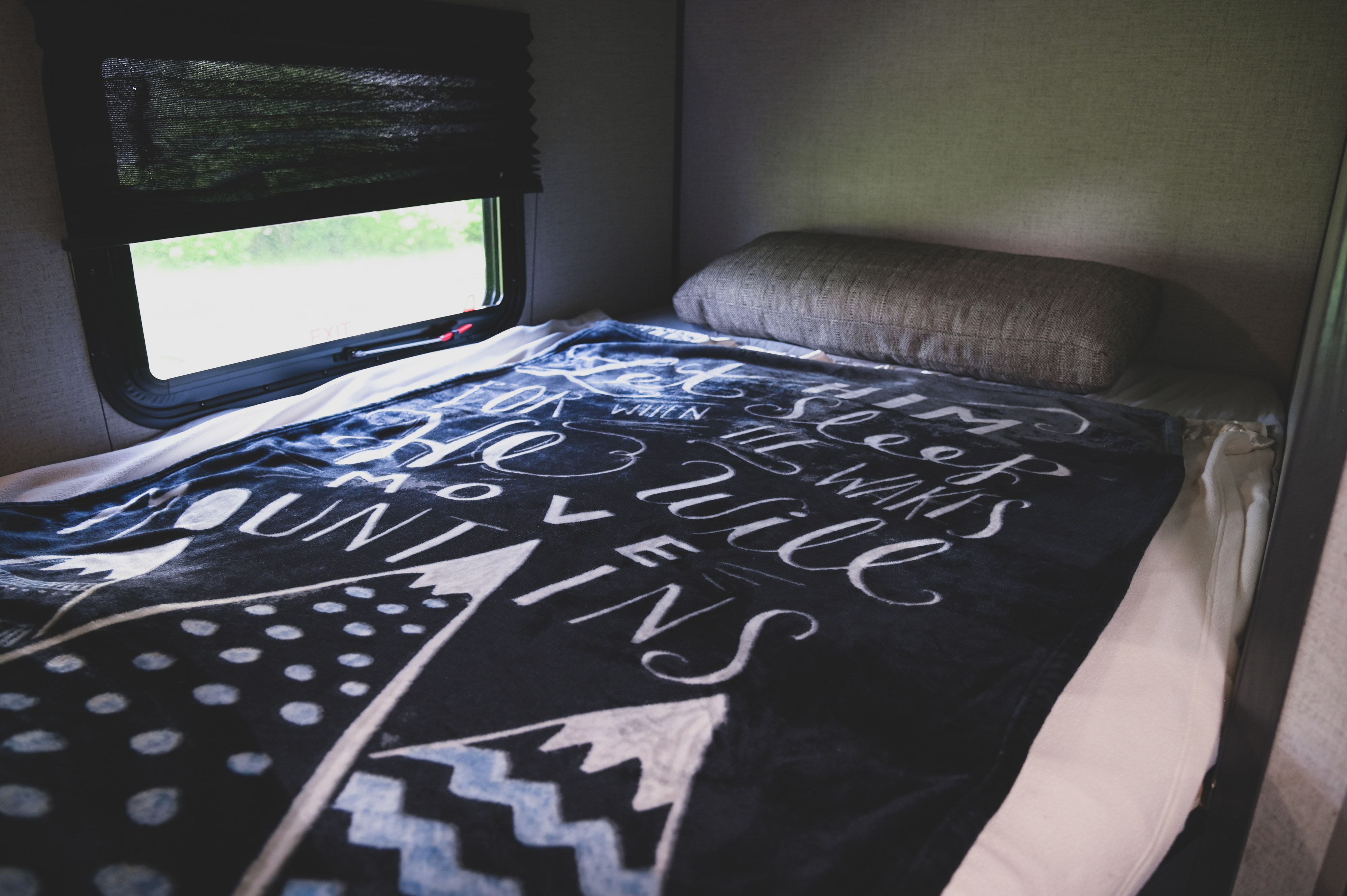 Inside an RV rental: a cozy bed with a decorative blanket, pillow, and window offering a glimpse of nature.. Aspen Trail LE 2021