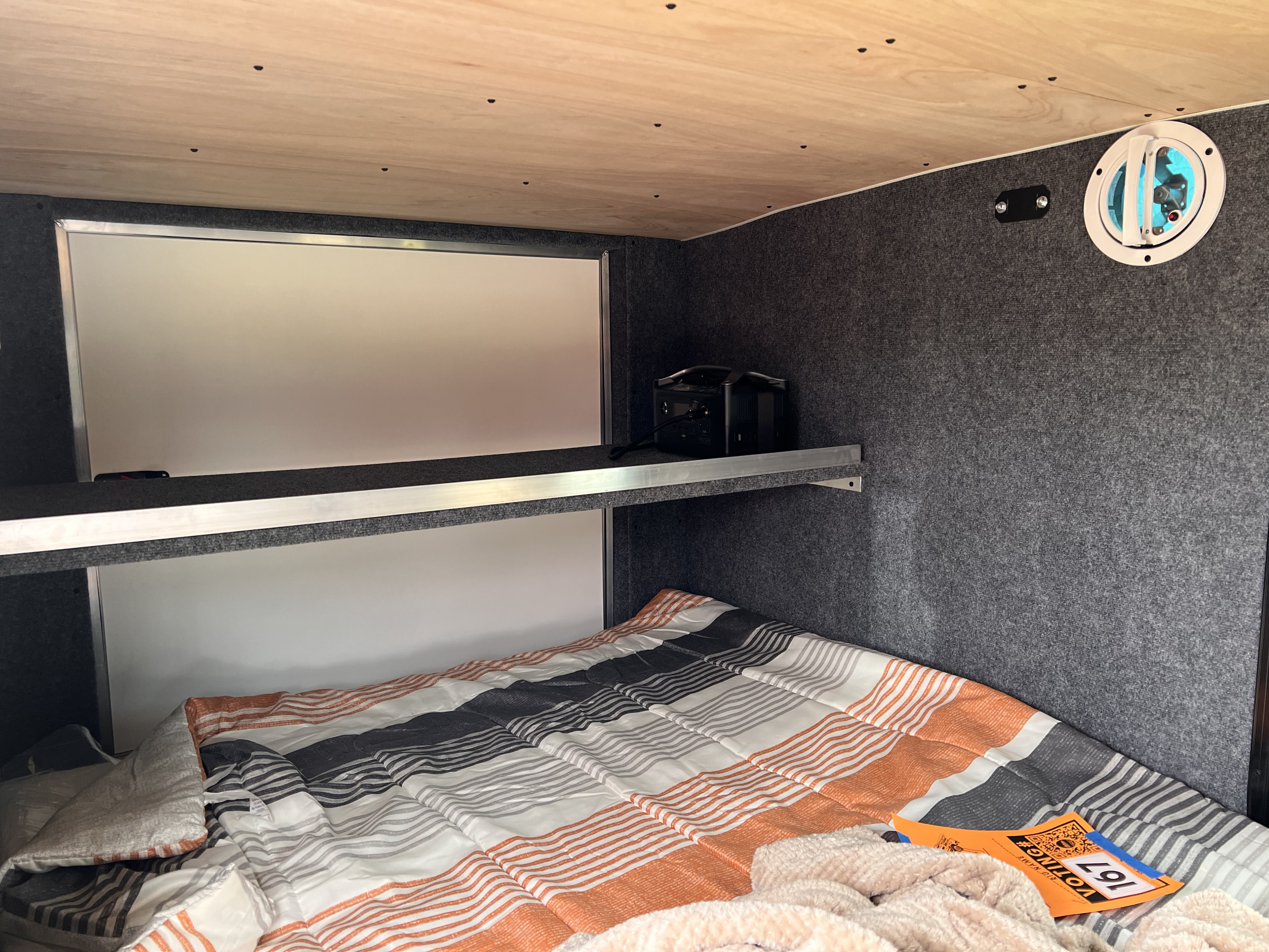 Cozy RV rental interior with a striped bed, overhead shelf holding a power station, and side wall vent for a comfortable sleep setup.. Sherpa trailers Big foot 2022