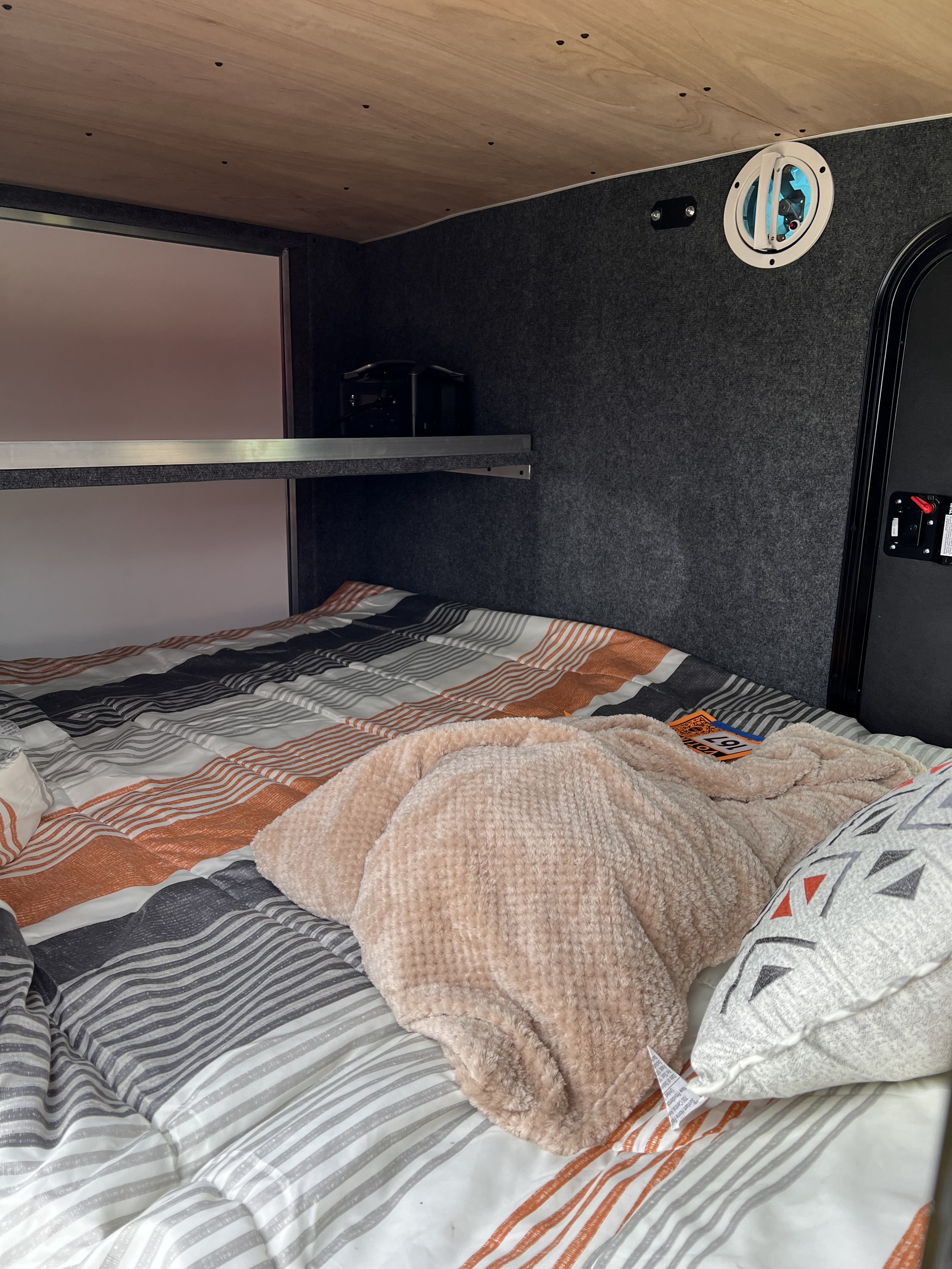 An inviting rv rental sleeping area, featuring a striped bedspread, soft blanket, wooden ceiling, and a practical shelf unit.. Sherpa trailers Big foot 2022