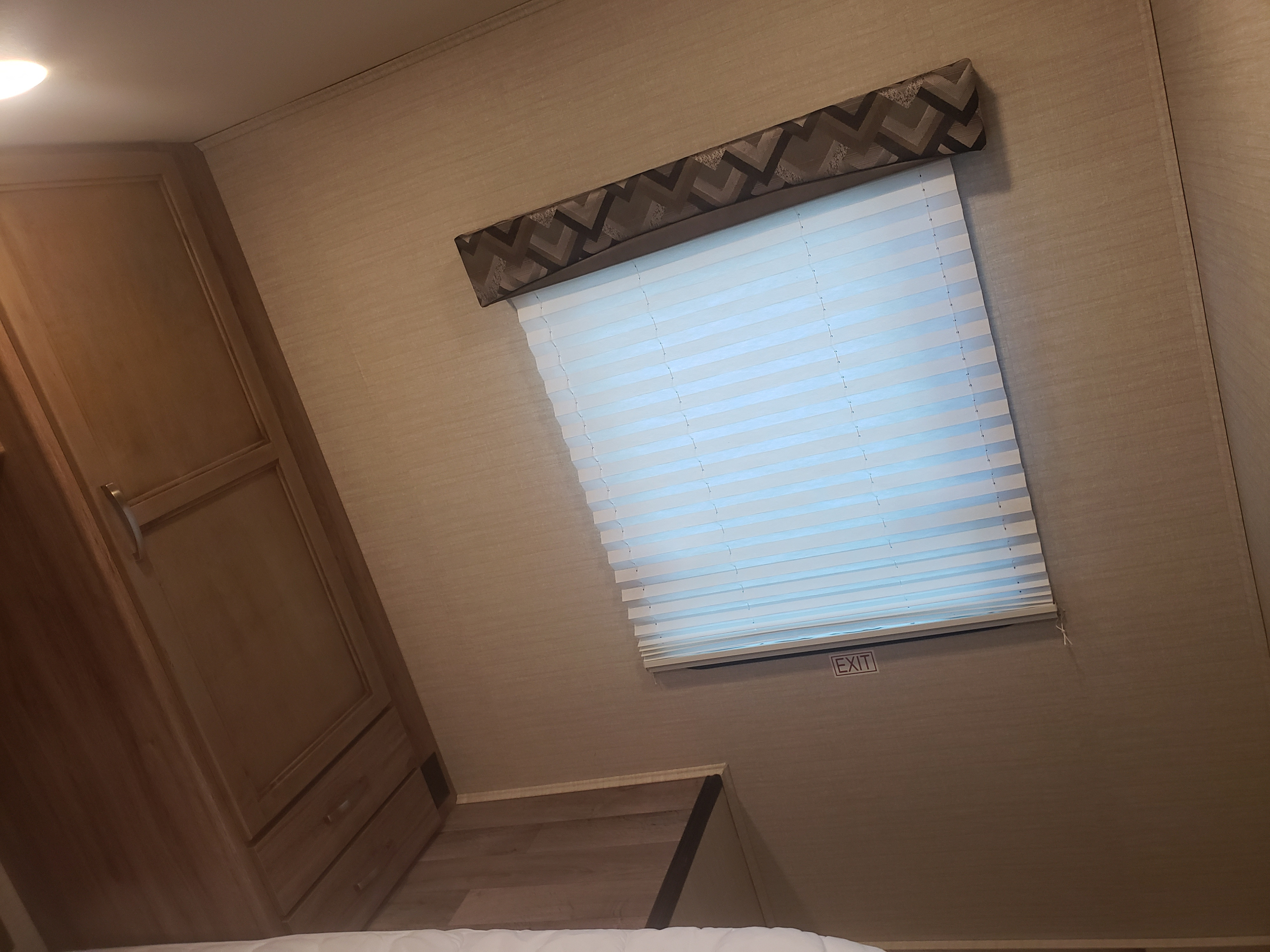 RV rental interior: A compact space with a window featuring white blinds and a patterned valance, a wooden cabinet, and an "EXIT" sign.. Jayco Redhawk SE 2019