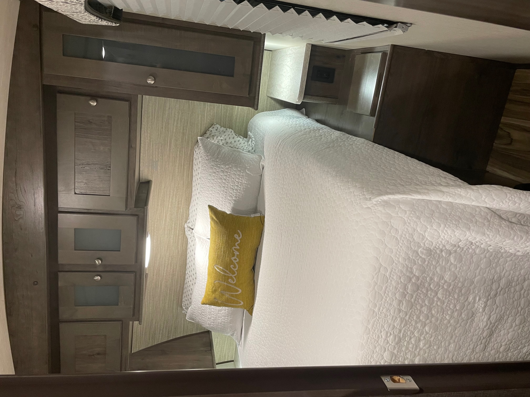 Inside a comfy RV rental bedroom: white quilted bed, 'Welcome' pillow, and dark wood cabinetry. Ideal for your next road trip.. Cruiser RV MPG Ultra Lite 2019