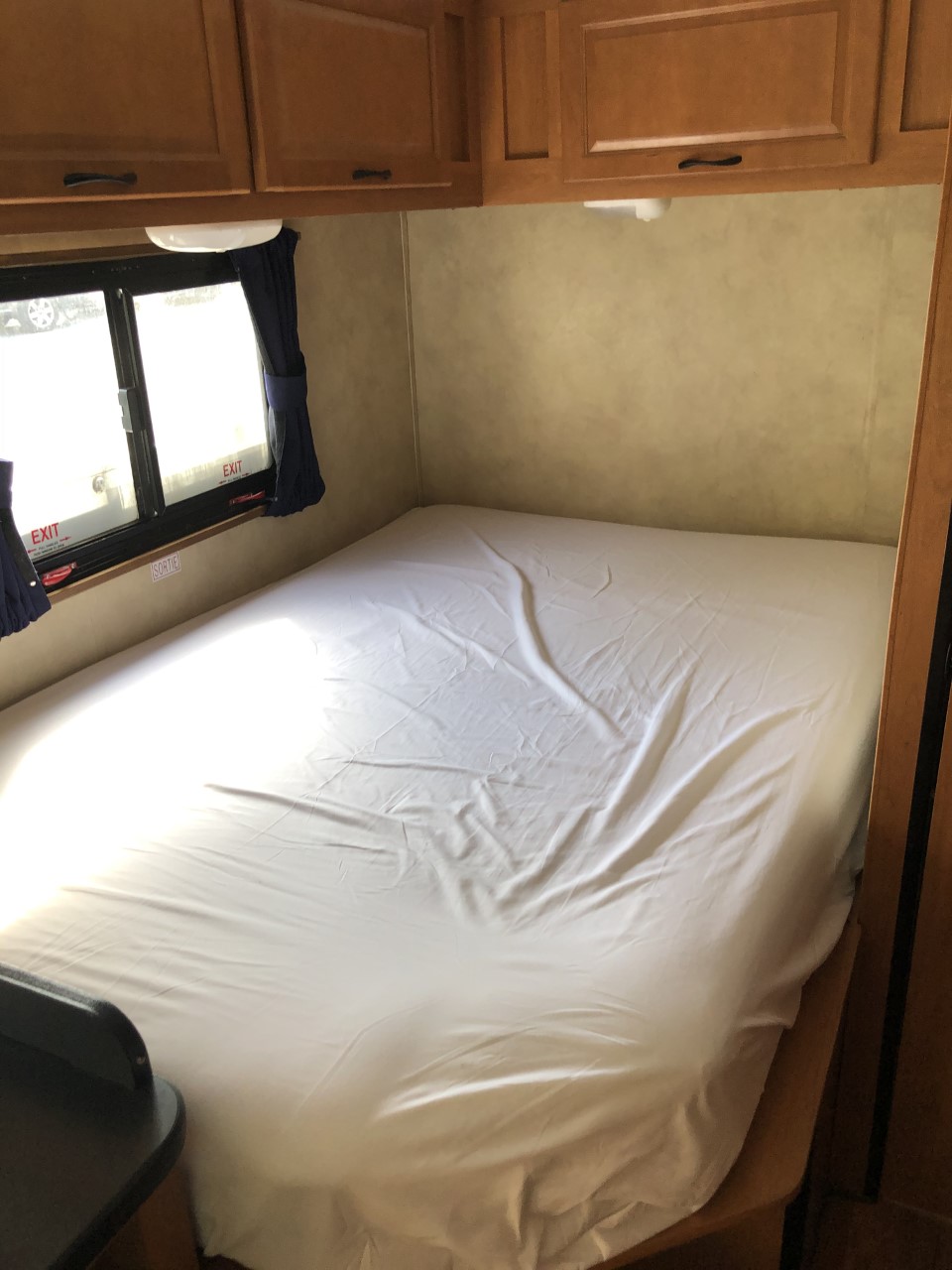 Inside an RV rental: a neatly made bed, overhead cabinets, and a window with visible 'EXIT' signs.. THOR MAJESTIC 23a 2018