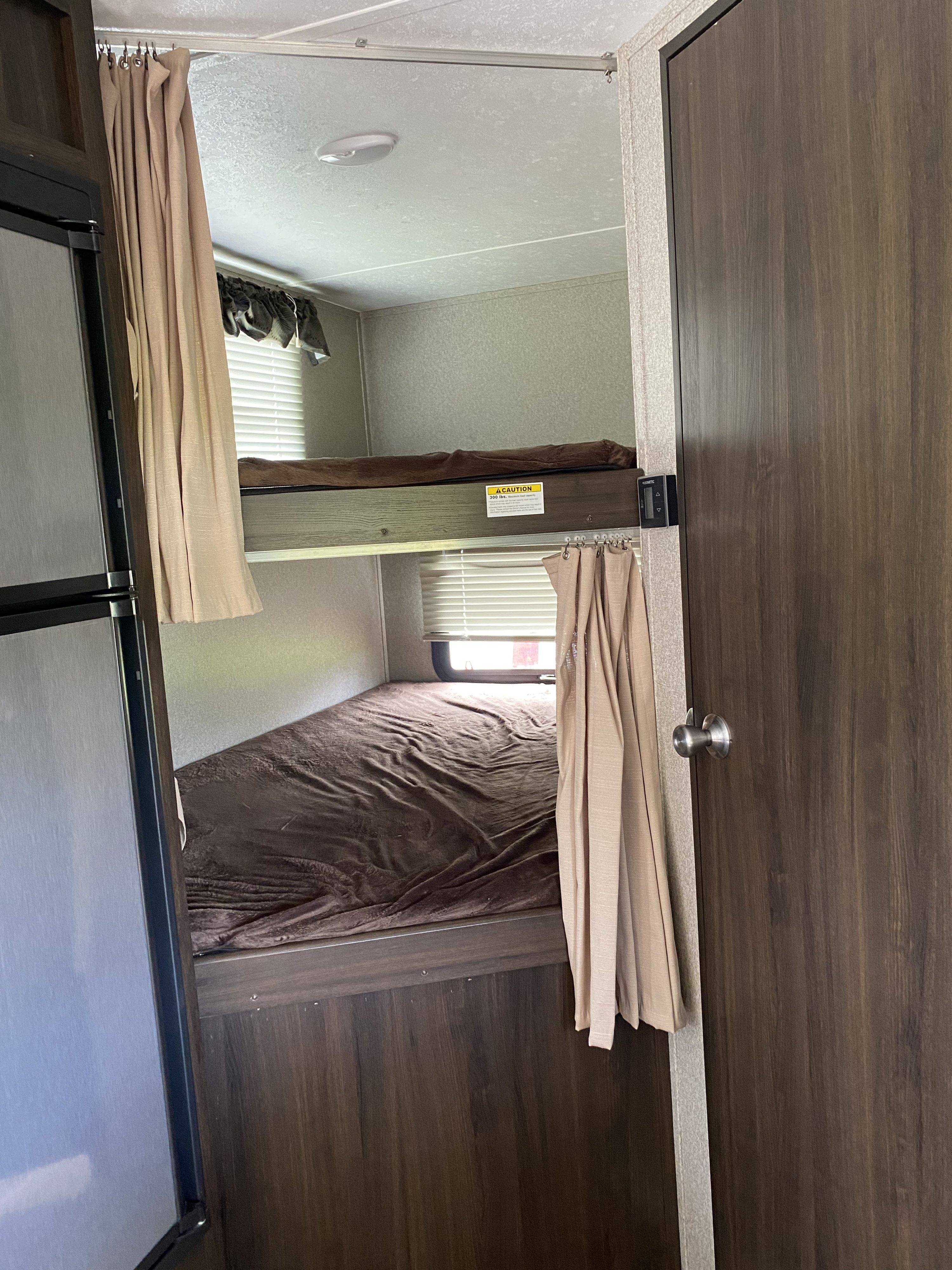 RV rental interior showing stacked bunk beds with brown blankets, privacy curtains, a fridge, and dark wood finishes.. Keystone RV Springdale 2018