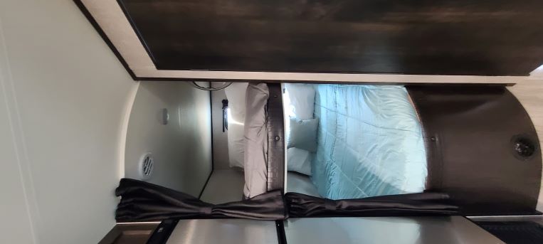 This RV rental's interior features a bed with blue bedding, a compact shower area, and dark overhead cabinetry.. Jayco Flight 242BHS 2022