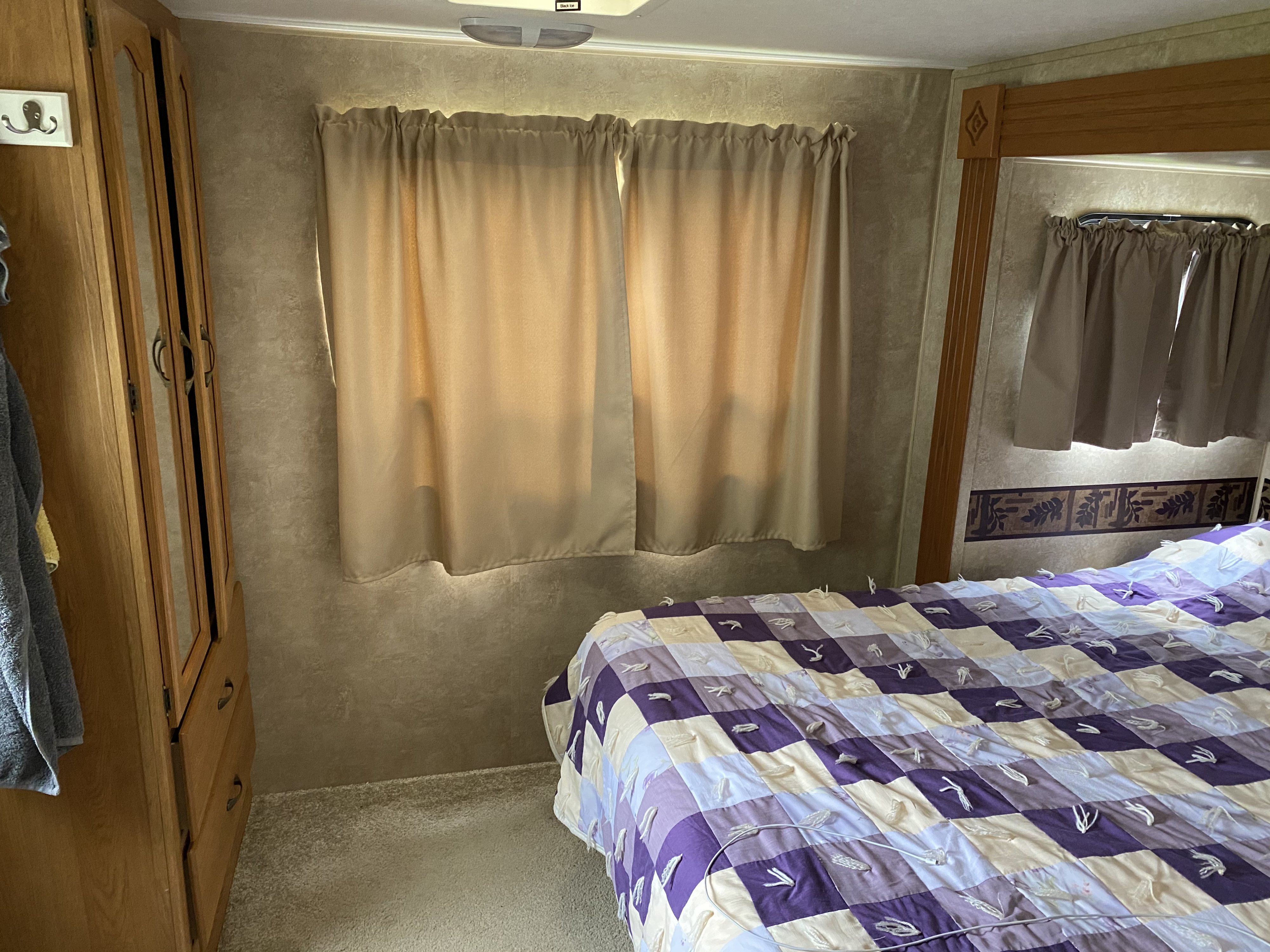 Cozy RV rental bedroom featuring a purple and white checkered quilt, light curtains, and a wooden wardrobe.. Keystone RV Springdale 2010