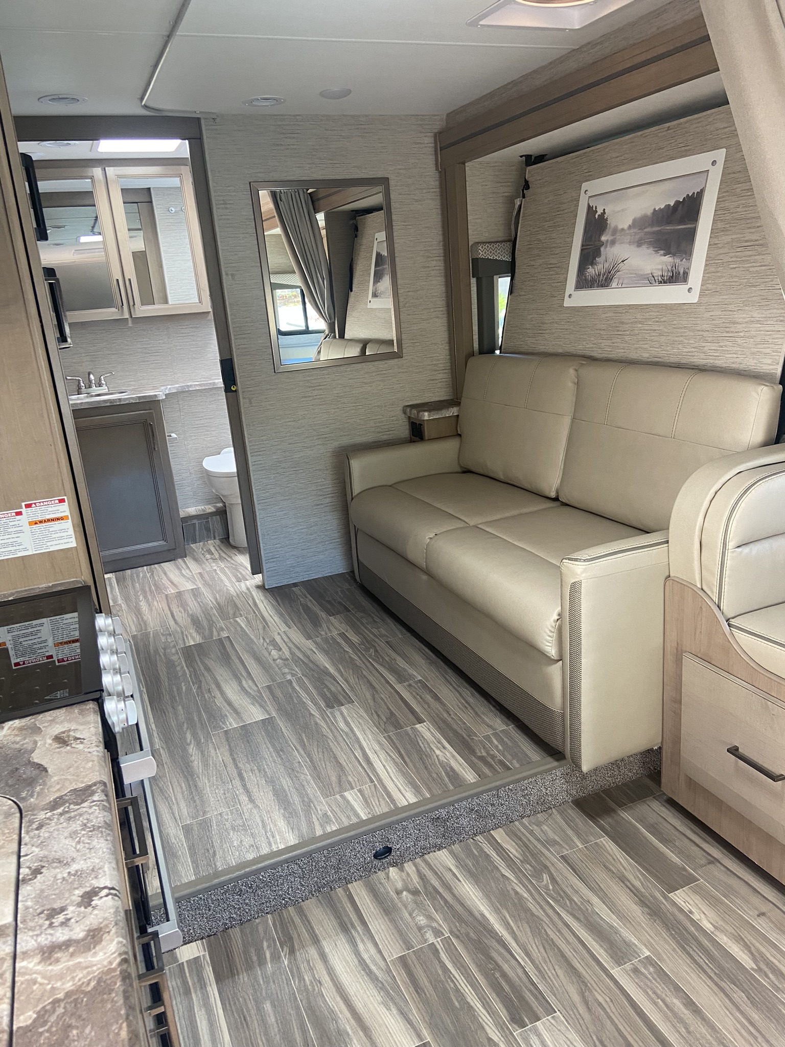 Modern RV rental interior with a light-colored sofa, wood-look flooring, and glimpses of the kitchen and bathroom.. Thor Motor Coach Four Winds 25M 2022