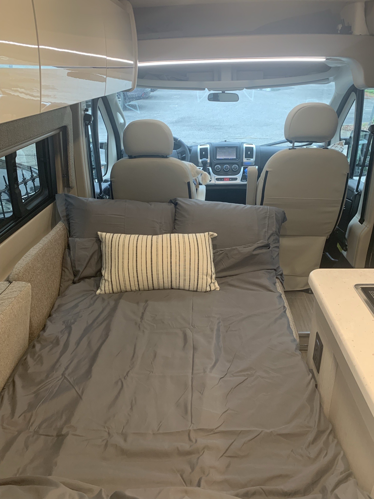 Cozy RV rental interior: Full-size bed with grey bedding, compact kitchen, and front cockpit of a camper van.. Thor Rize 18M 2022