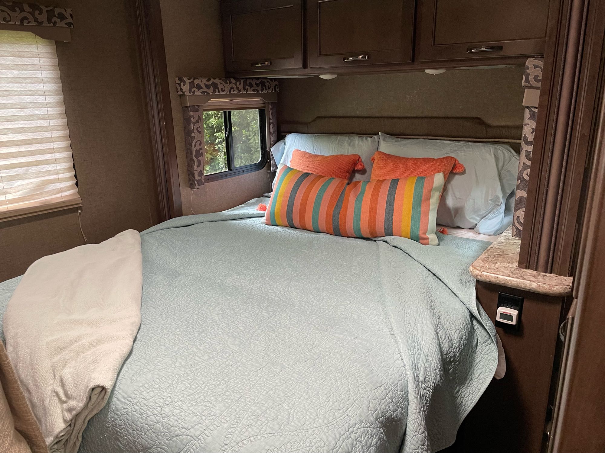 Cozy RV rental bedroom with light blue quilted bedding, bright striped and solid orange pillows, and overhead cabinets.. Thor Four Winds 2018