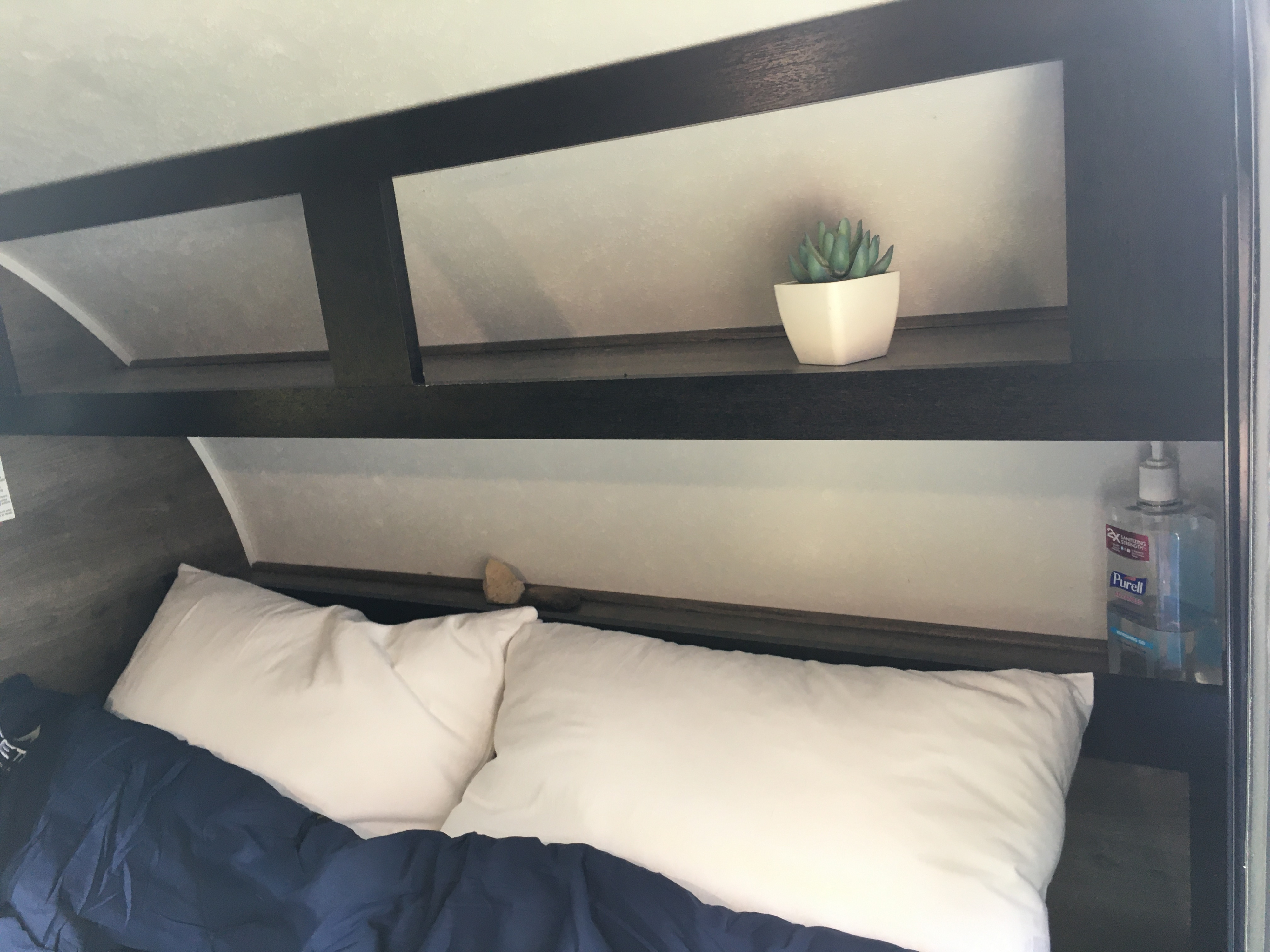 An RV rental's cozy bedroom features a bed with white pillows & blue blanket, plus overhead storage with a plant & Purell.. Braxton Creek Bushwhacker 2021
