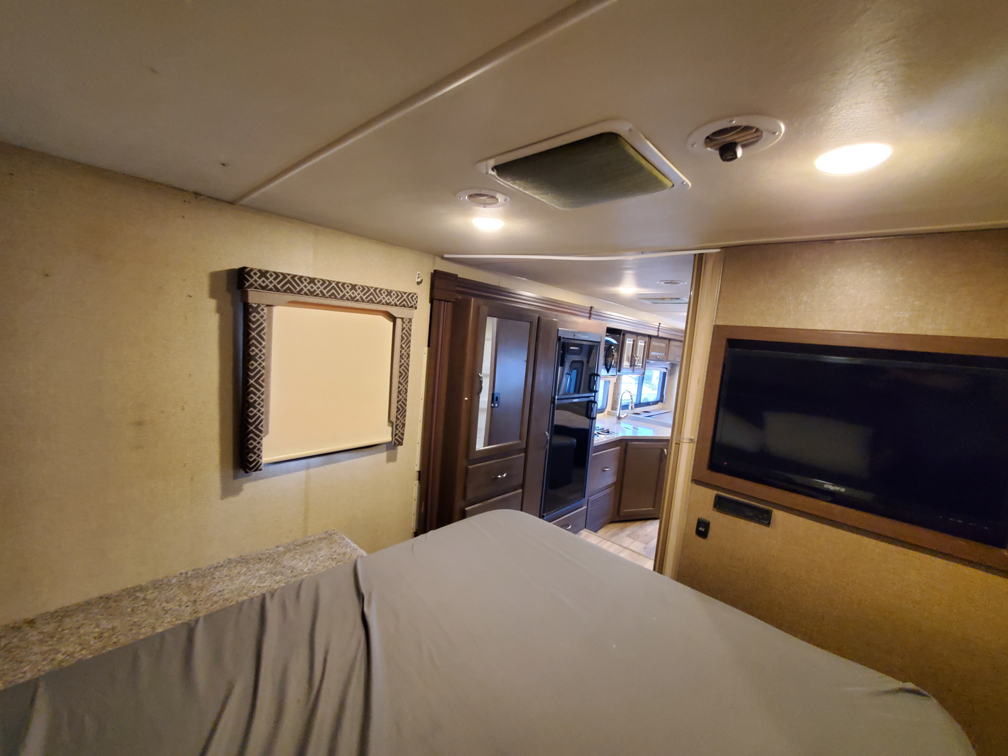 Bedroom of an RV rental, featuring a bed, wardrobe, and mounted TV. Connects to a full kitchen area visible beyond.. Thor Motor Coach Four Winds 2019