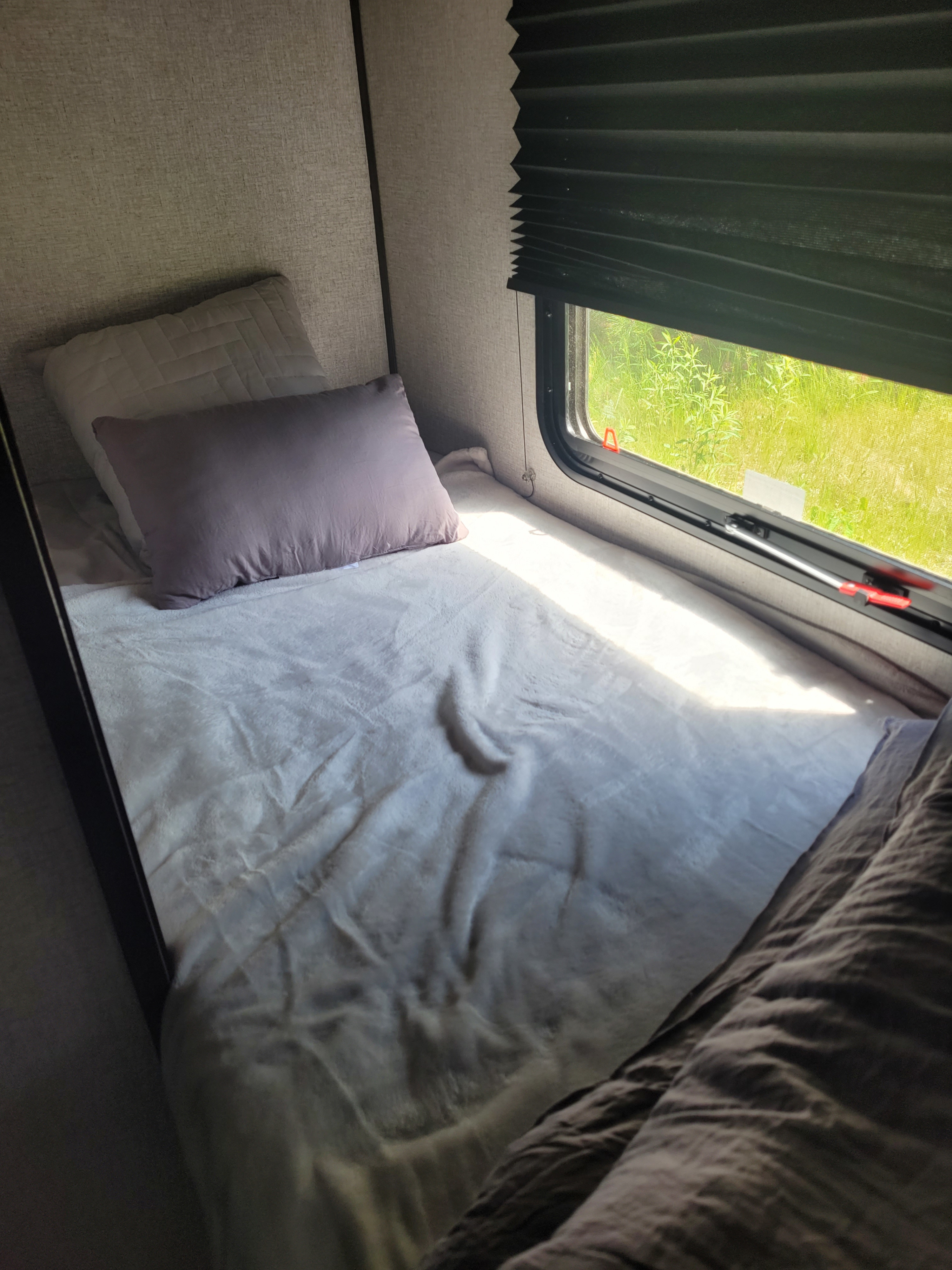 Cozy single bed in an RV rental with light gray blanket, pillows, and bright sun peeking through the window at green scenery.. Dutchmen Coleman Lantern LT 2021
