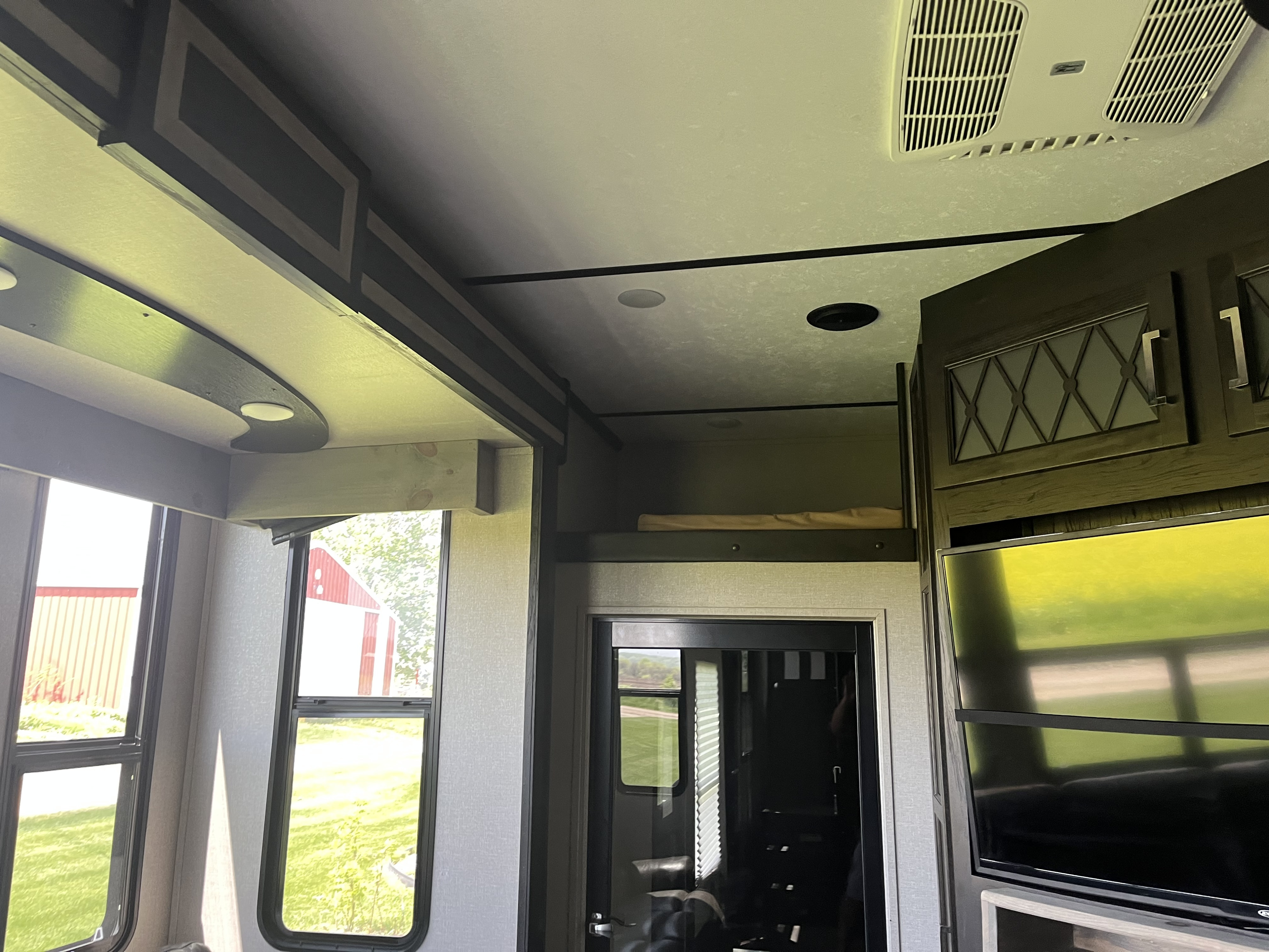 This RV rental interior showcases its ceiling with lights and AC, large windows overlooking a red barn, and a cozy upper bunk with TV.. Keystone Fuzion 2018