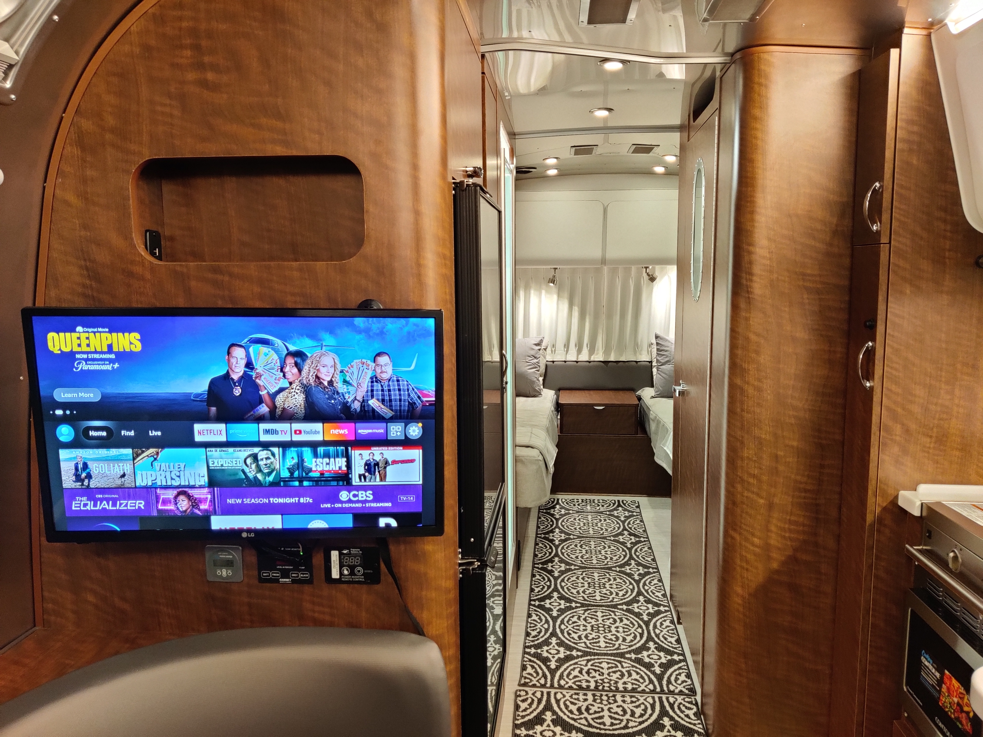 Cozy RV rental interior: TV displaying "Queenpins," controls, and a hallway leading to a bedroom with twin beds.. Airstream Globetrotter 2021