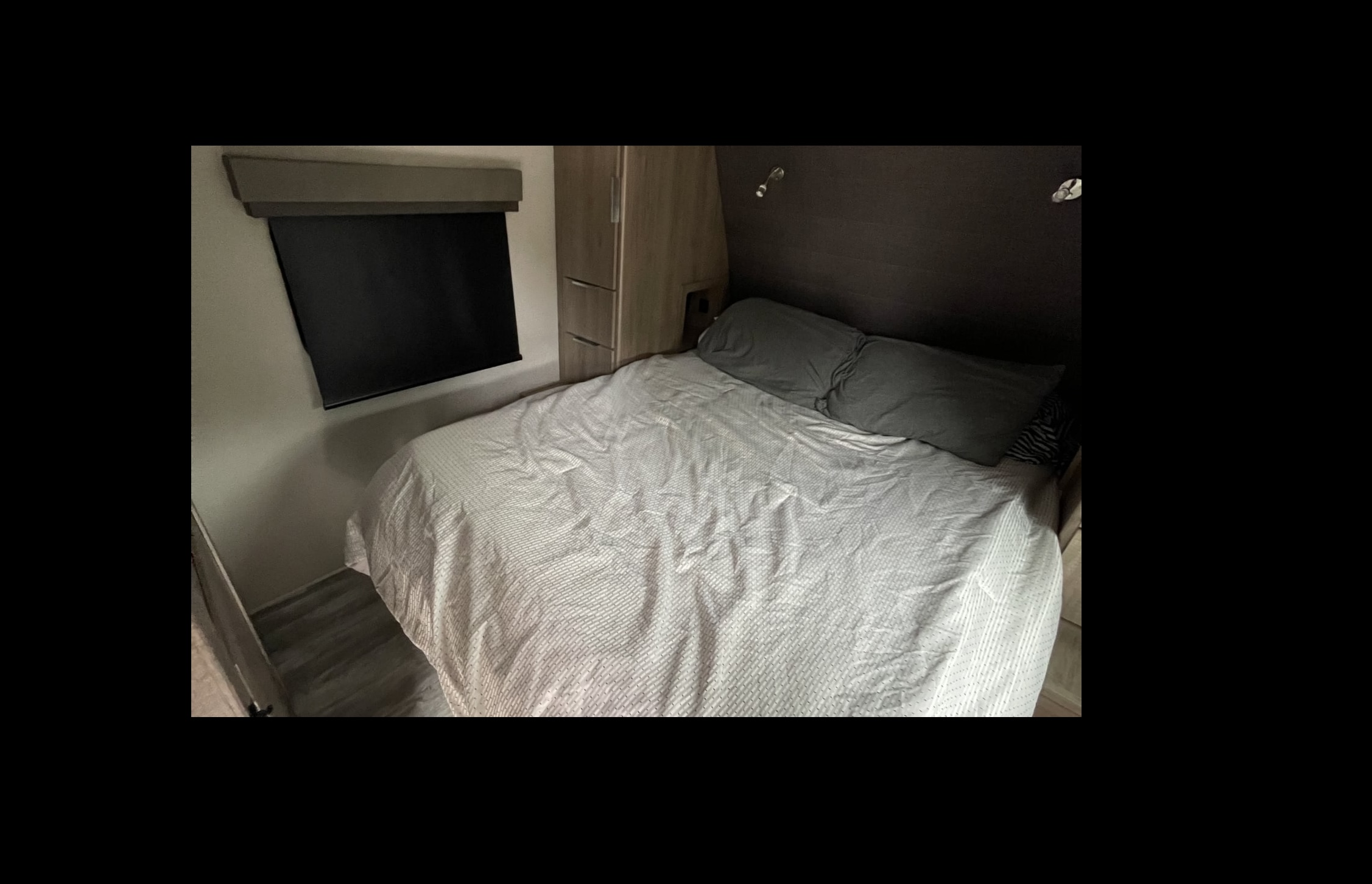 Inside a cozy rv rental: A compact bedroom features a bed with a white textured duvet, grey pillows, window with blind, and wood storage.. Grand Design Imagine XLS 2021