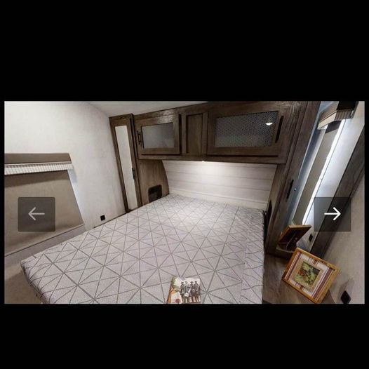 This RV rental interior features a cozy bedroom with a patterned bed, wood cabinets, and a side window, perfect for travel.. Forest River Wildwood 2021