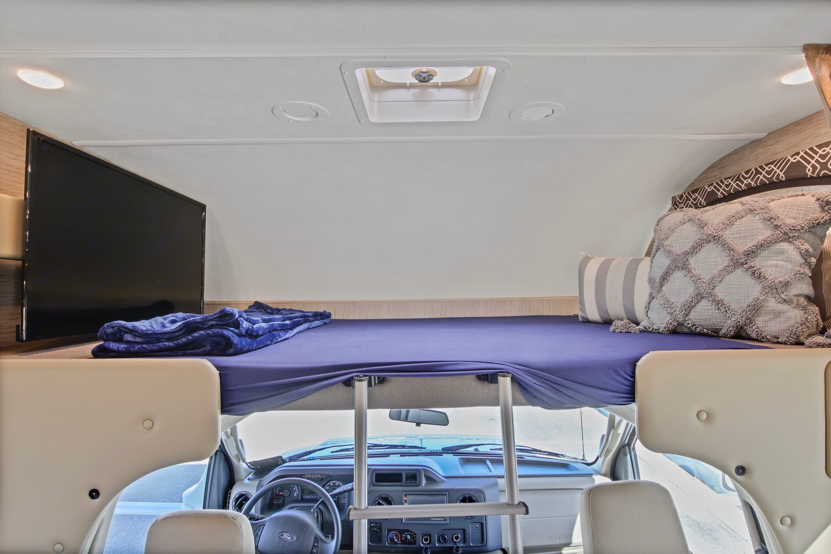 Comfortable over-cab sleeping area in an RV rental, featuring a TV, bedding, and a view into the cockpit.. Thor Chateau 2021