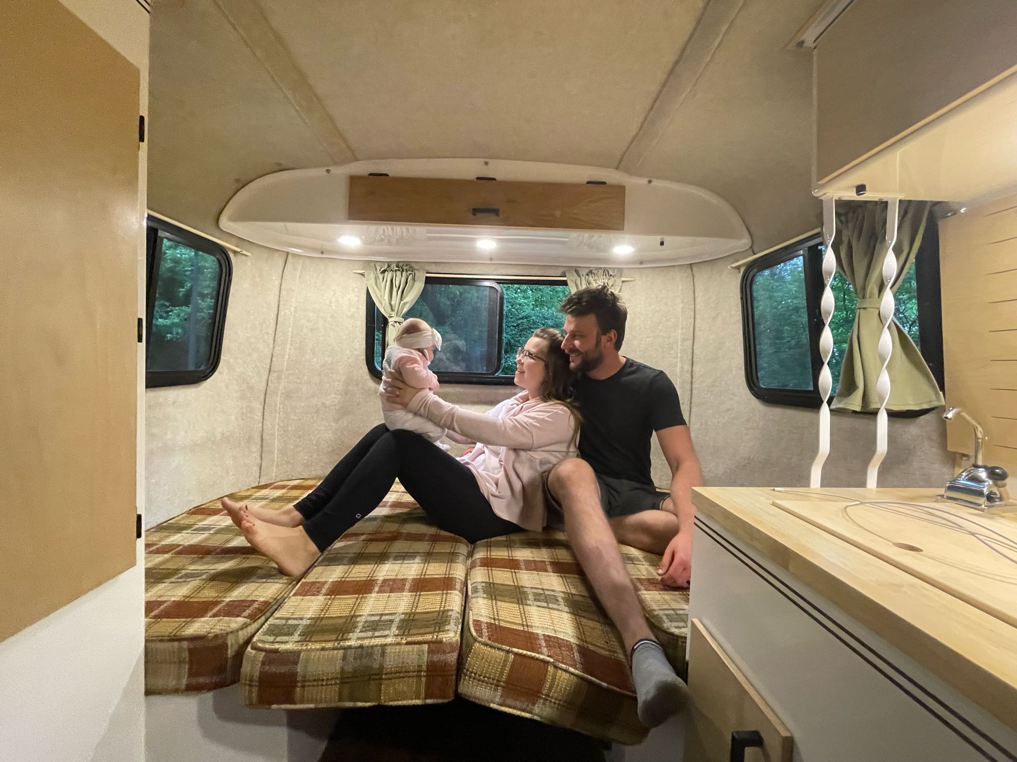 Family enjoying cozy moments in their RV rental. Parents & baby relax on a plaid bed, surrounded by nature views.. Scamp Scamp Trailer 2007