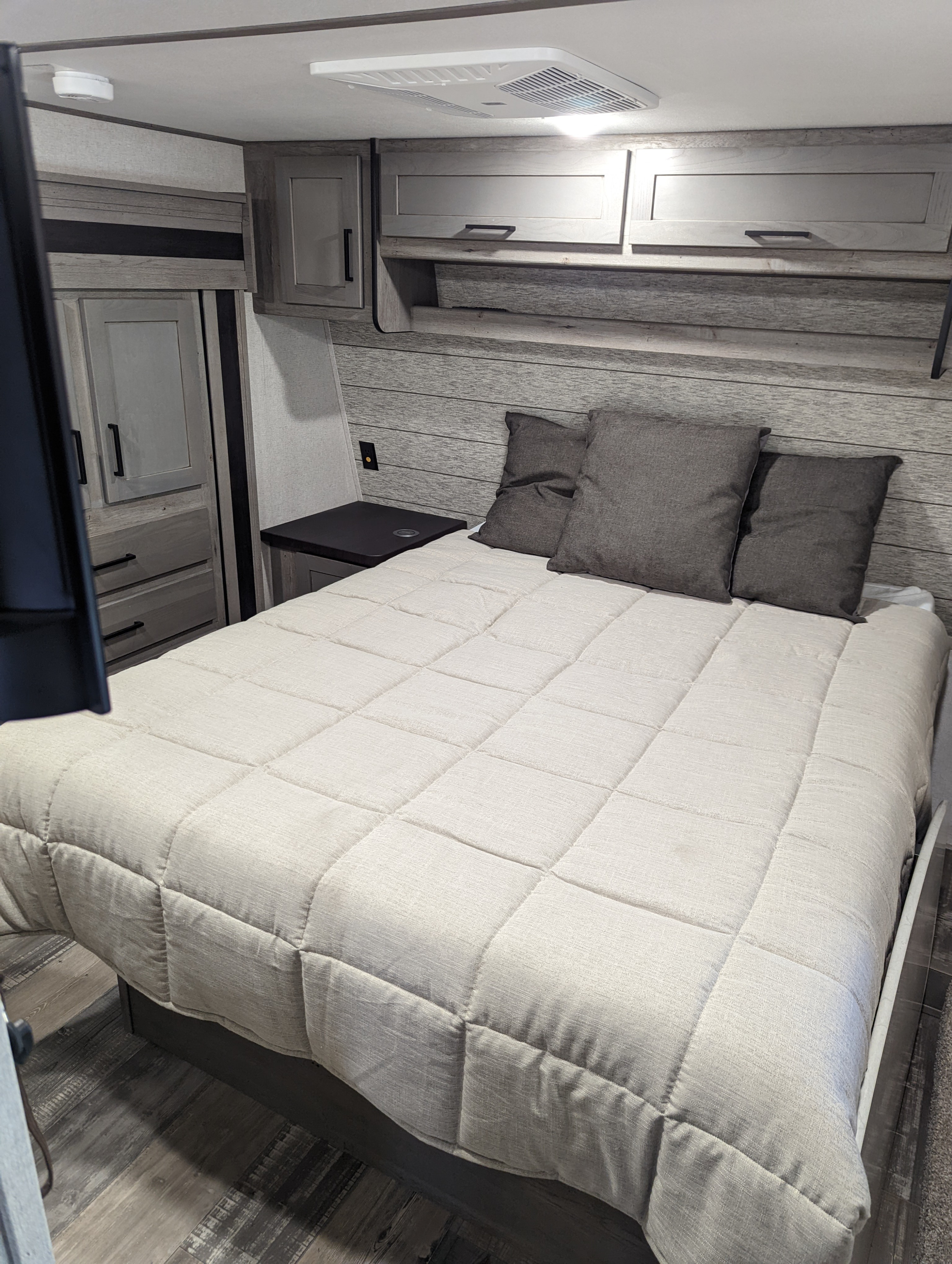 Interior of an RV rental bedroom: light quilt on bed, gray pillows, wood-paneled walls, and overhead cabinets for storage.. Keystone Raptor 2021