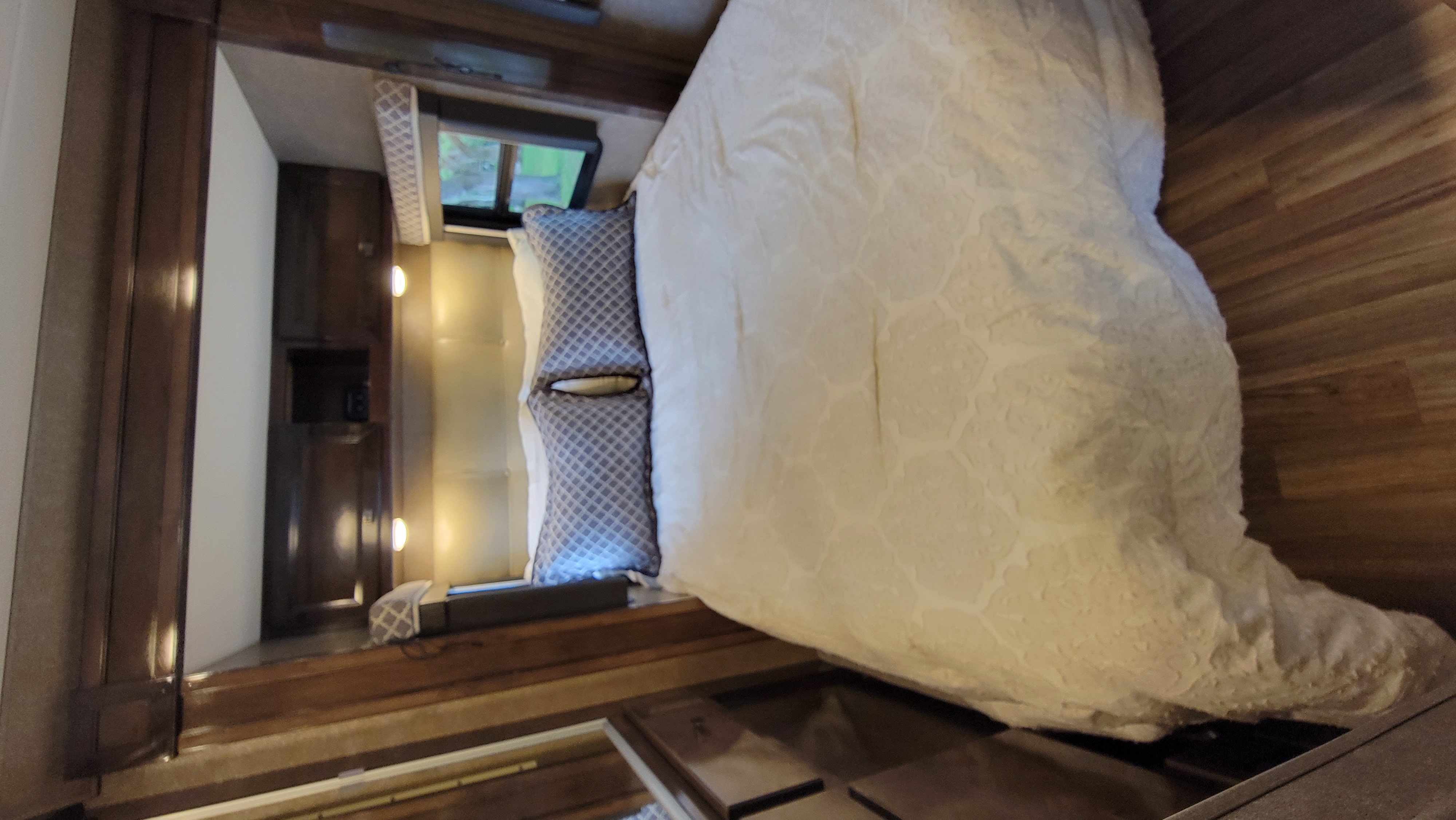 Cozy RV rental bedroom featuring a patterned comforter, wood floors, dark wood cabinetry, and window view.. Thor Synergy 2019