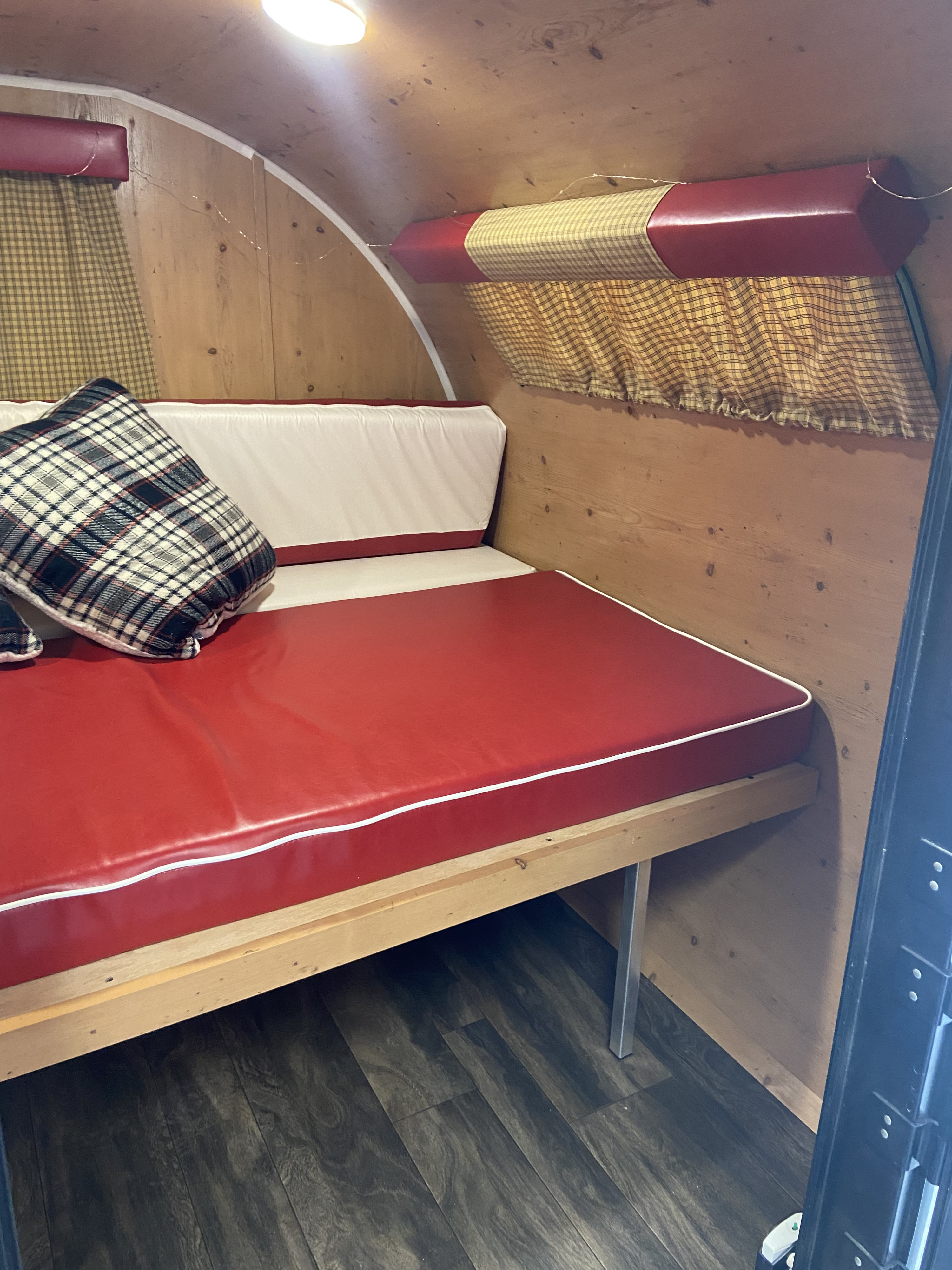 Cozy RV rental interior with a red and white upholstered bench bed, plaid pillow, wooden walls, and gingham curtains.. Sunset Park RV SunRay 2020