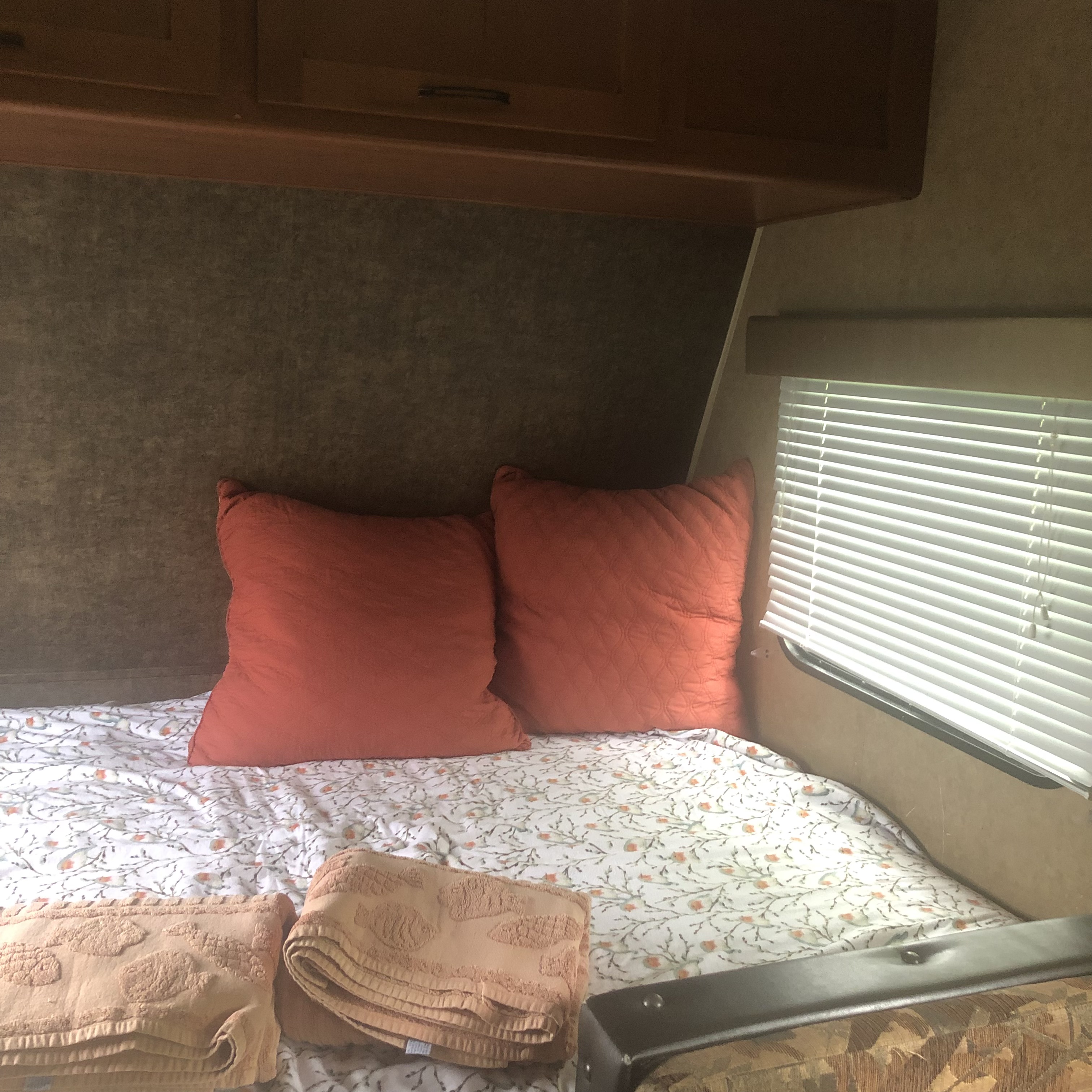 RV rental bedroom with a bed, floral sheets, orange pillows, folded towels, overhead cabinets, and a window with blinds.. Forest River Cherokee 2014