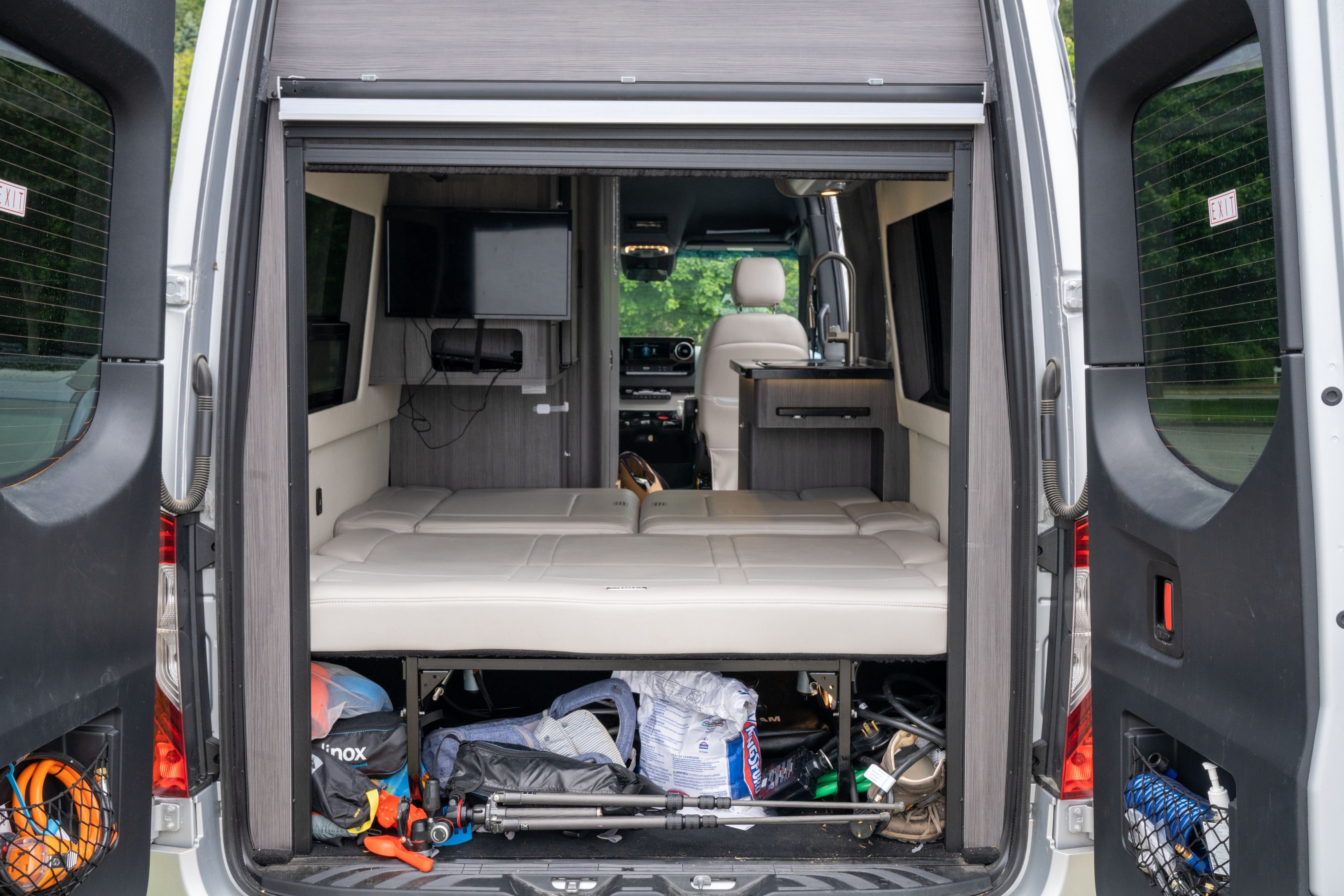 RV rental interior: A modern camper van with a versatile bed, under-bed storage, TV, and compact kitchen, seen from open rear doors.. Airstream Interstate 2020