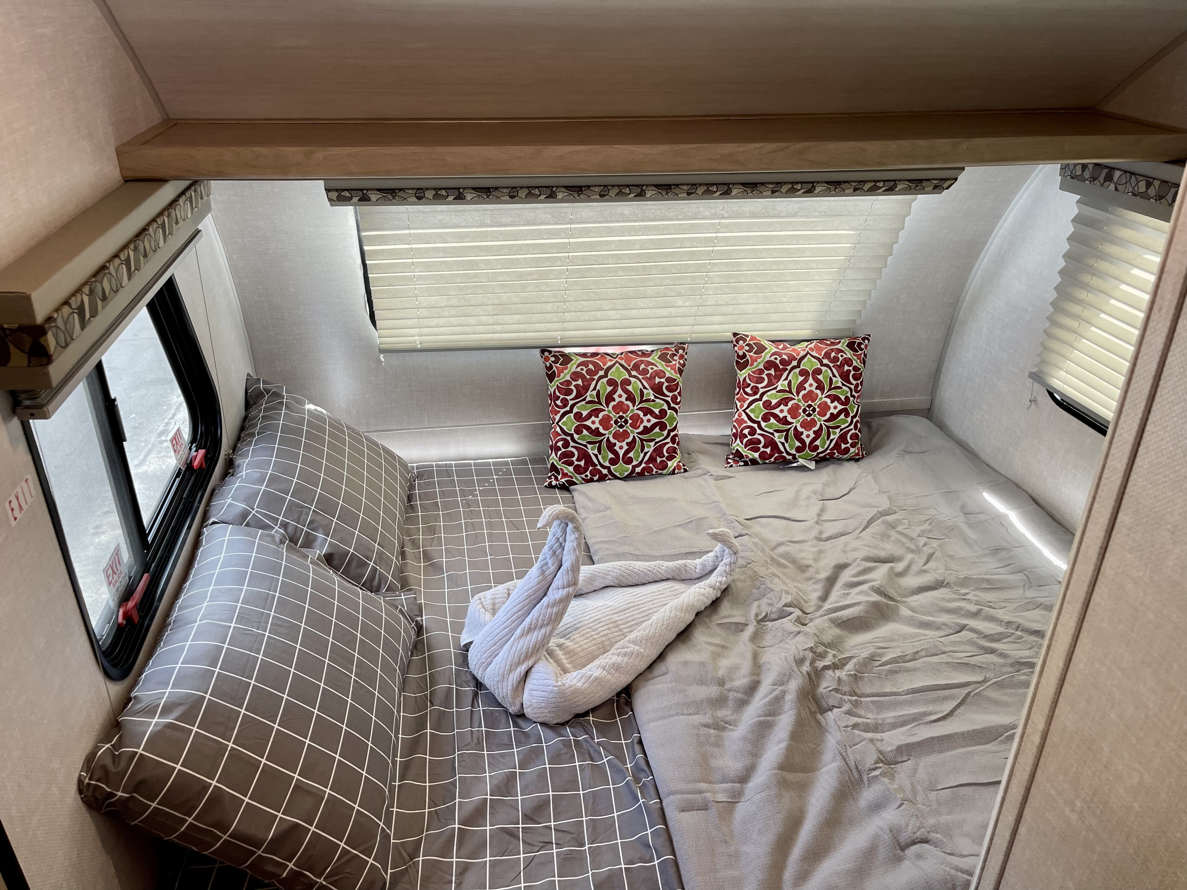 Cozy RV rental bedroom features a bed with grey plaid and plain bedding, decorative pillows, and a swan-folded towel. An emergency exit window is visible.. R-Pod R-Pod Trailer 2021