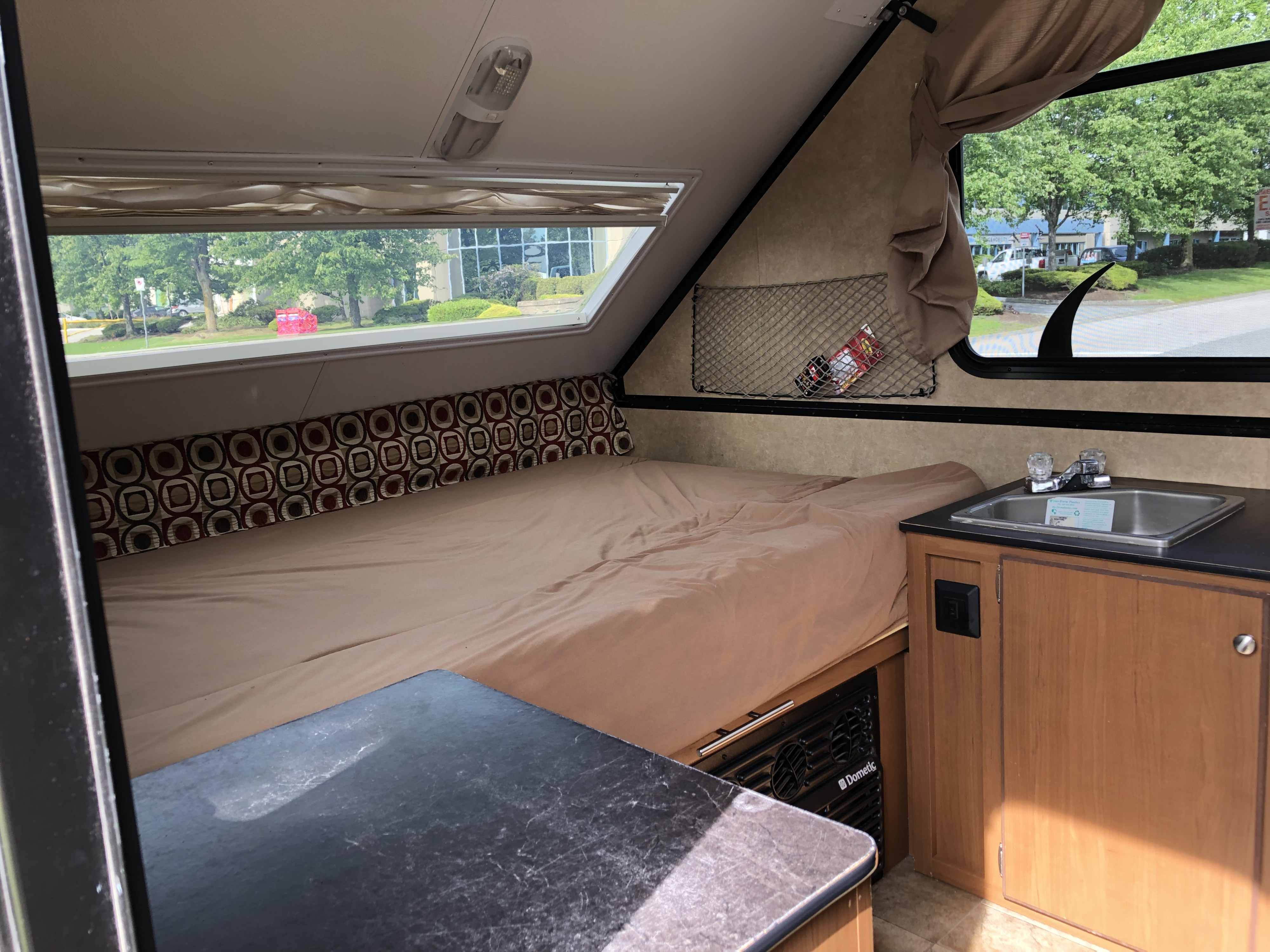 Interior of an RV rental showing a made bed, compact sink, and Dometic unit, with large windows overlooking trees and buildings.. Starcraft Comet 2015