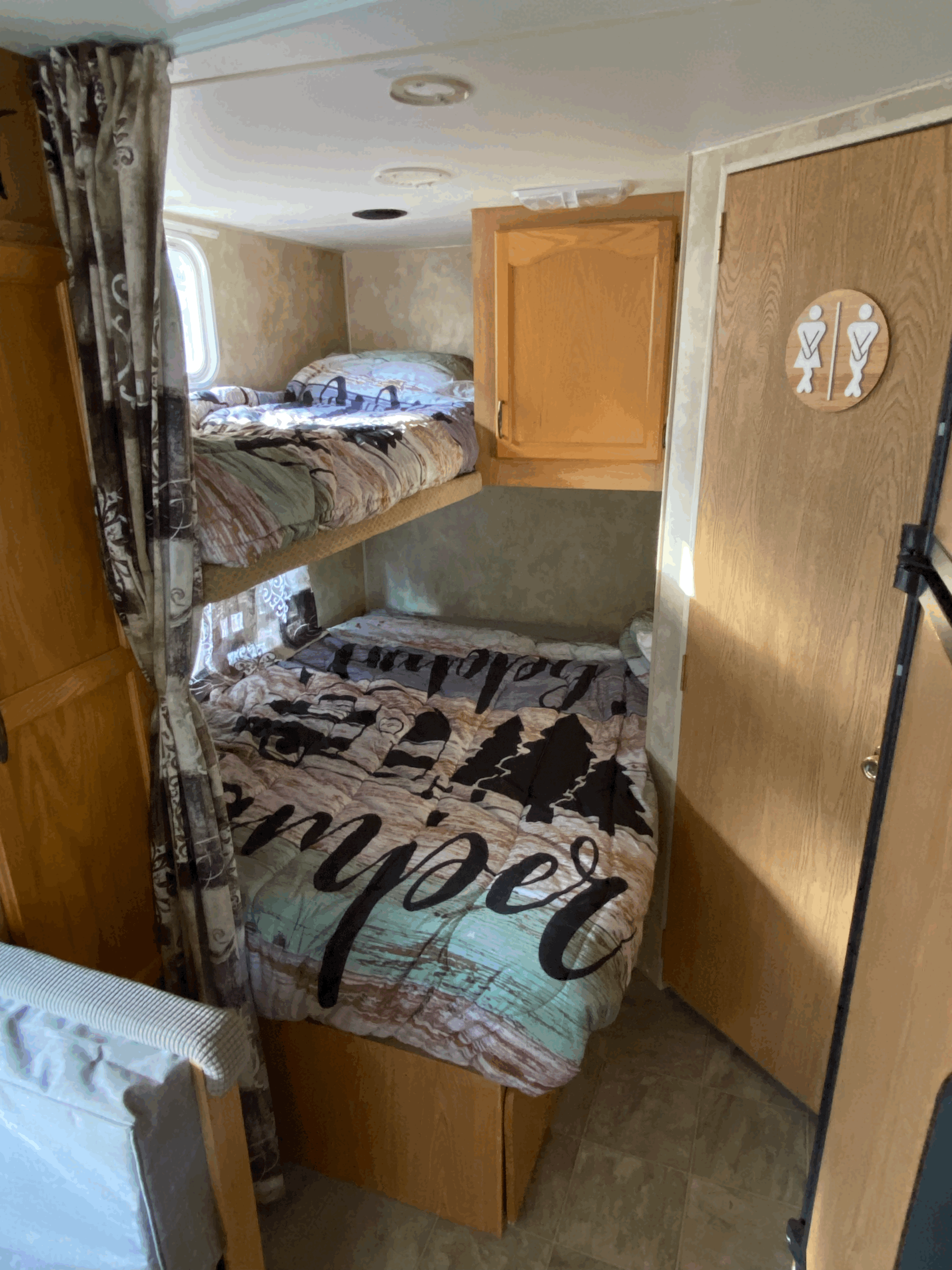 Inside an RV rental: built-in bunk beds with "Happy Camper" bedding, a curtain, and a restroom door with symbols.. Fleetwood Pioneer 2007