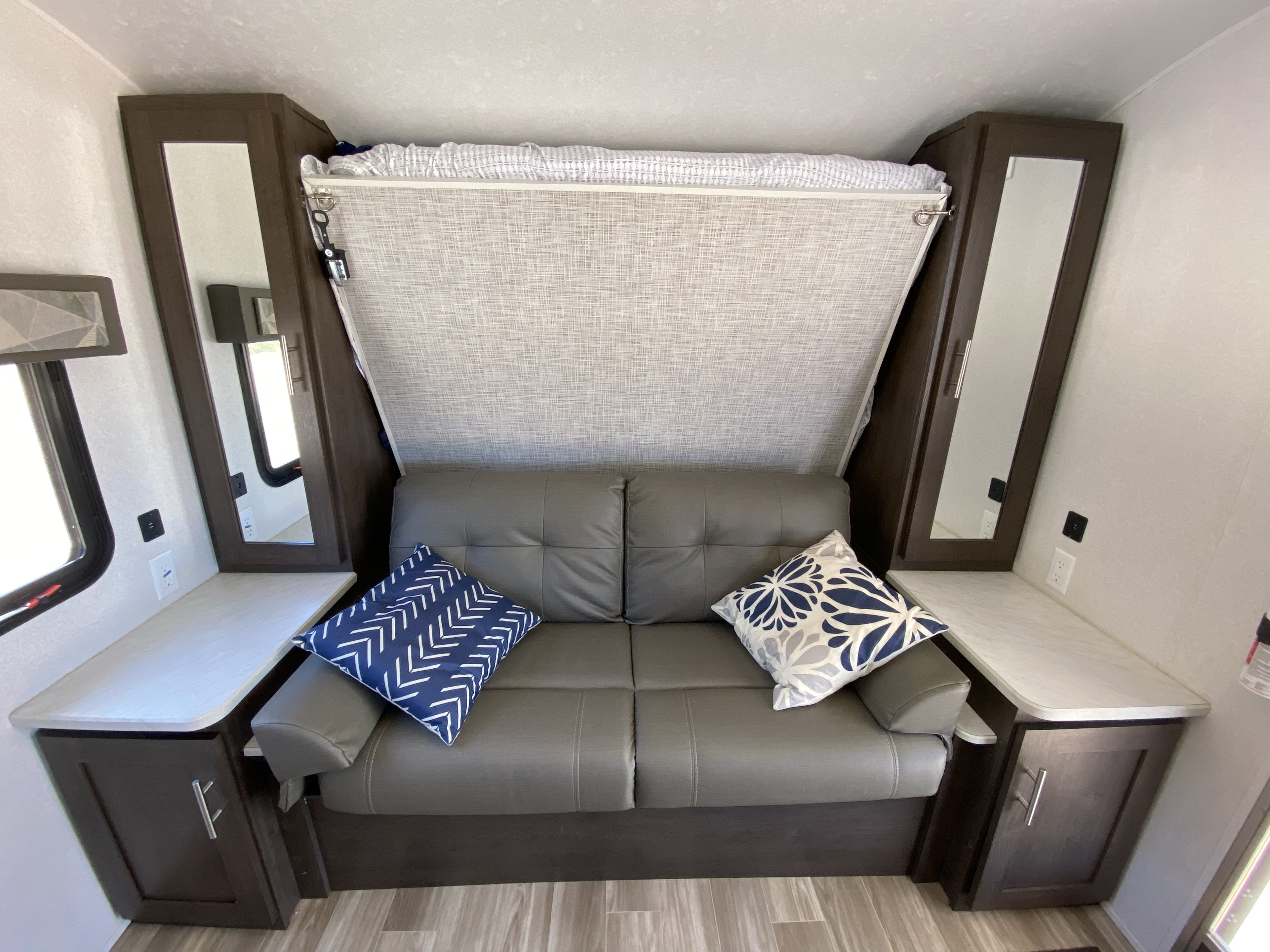 Bright RV rental interior featuring a grey convertible sofa, overhead bunk, and mirrored storage cabinets.. Forest River Wildwood X-Lite 2019
