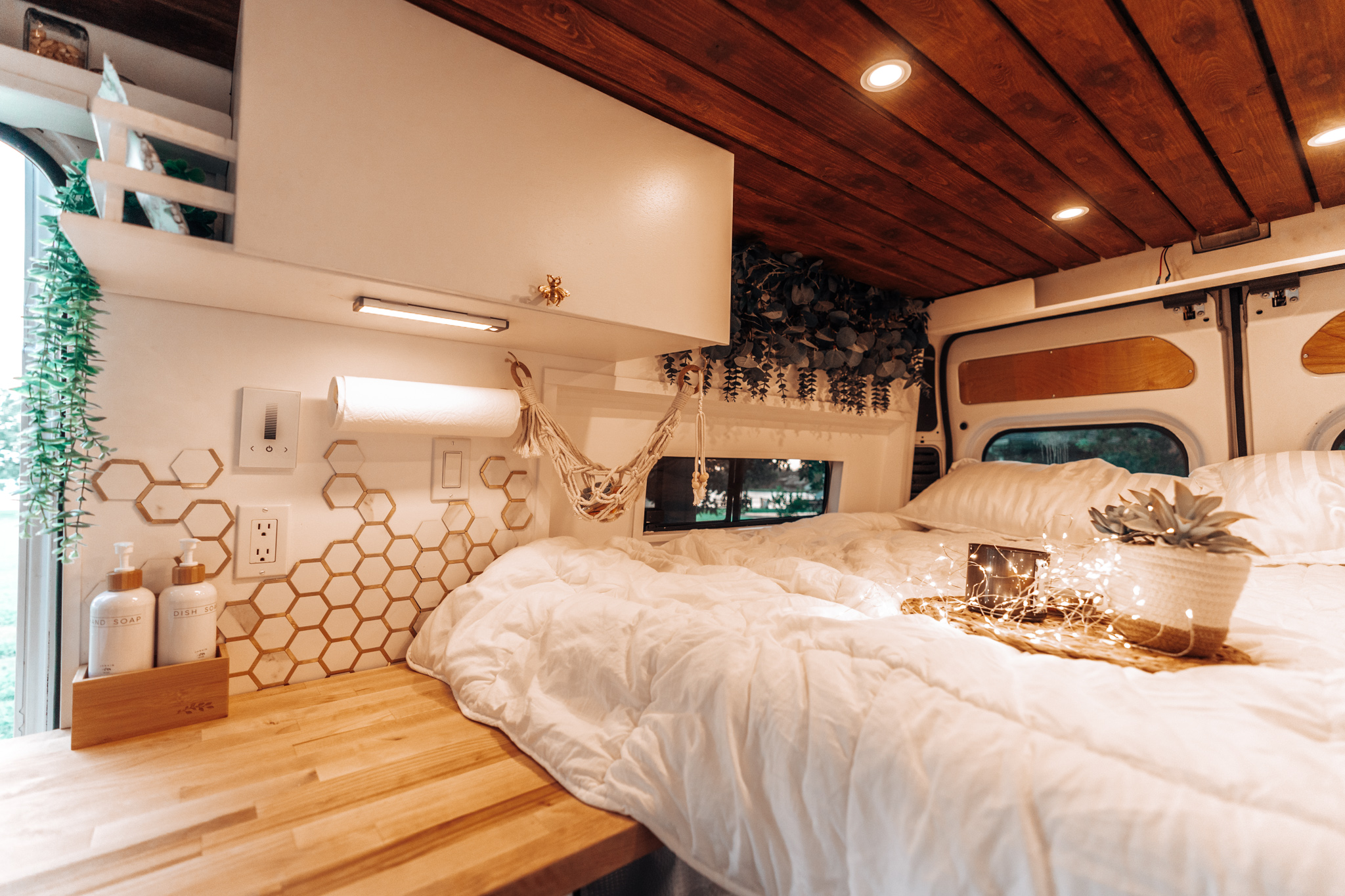 A bright rv rental interior with a comfy bed, wooden ceiling & counter, white cabinets, and modern honeycomb backsplash.. RAM Promaster 2015