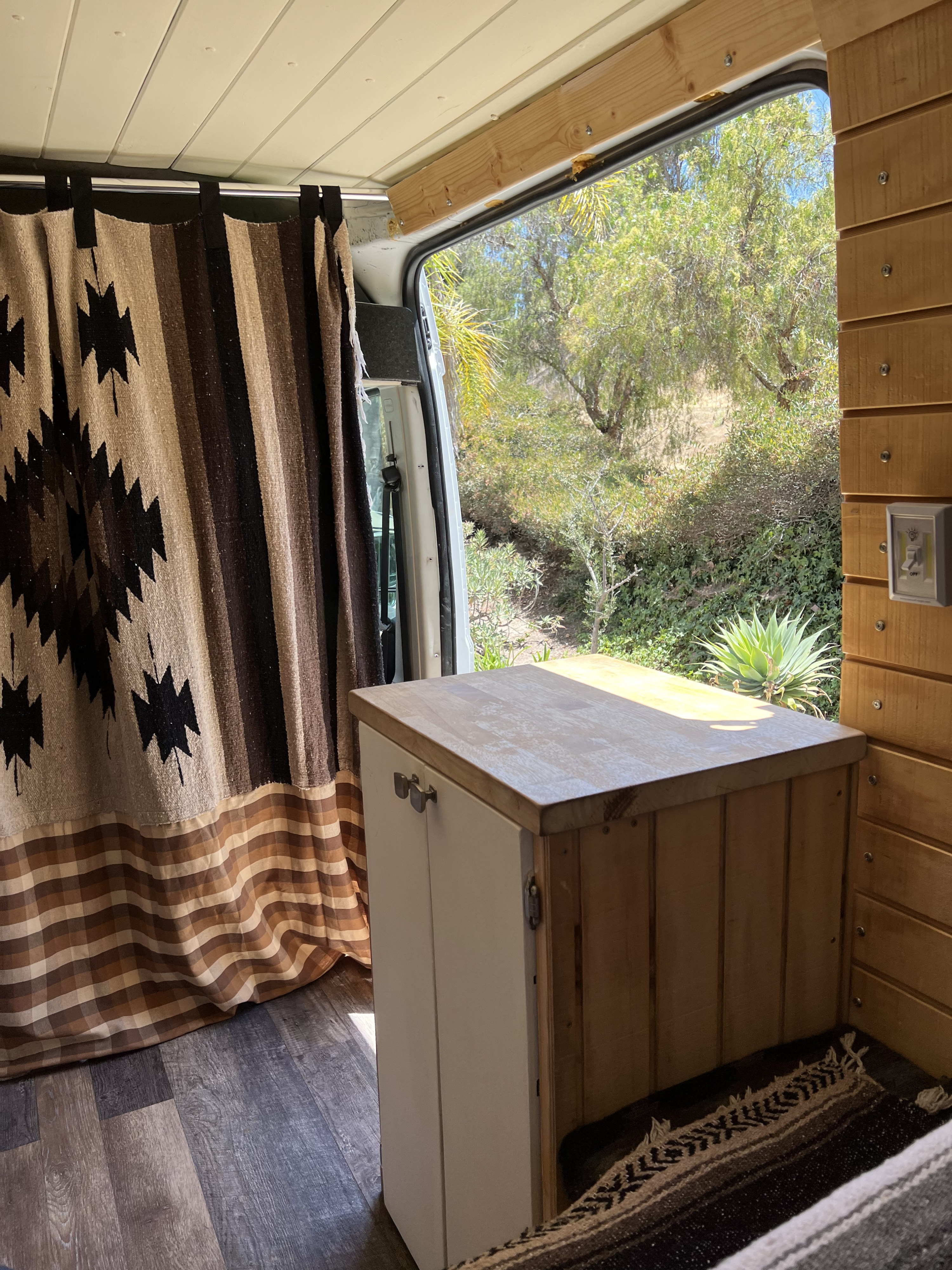 Cozy RV rental interior with wood details, Southwestern curtain, and a bright outdoor view of trees and a succulent from the open door.. Dodge Ram Promaster 2500 2017