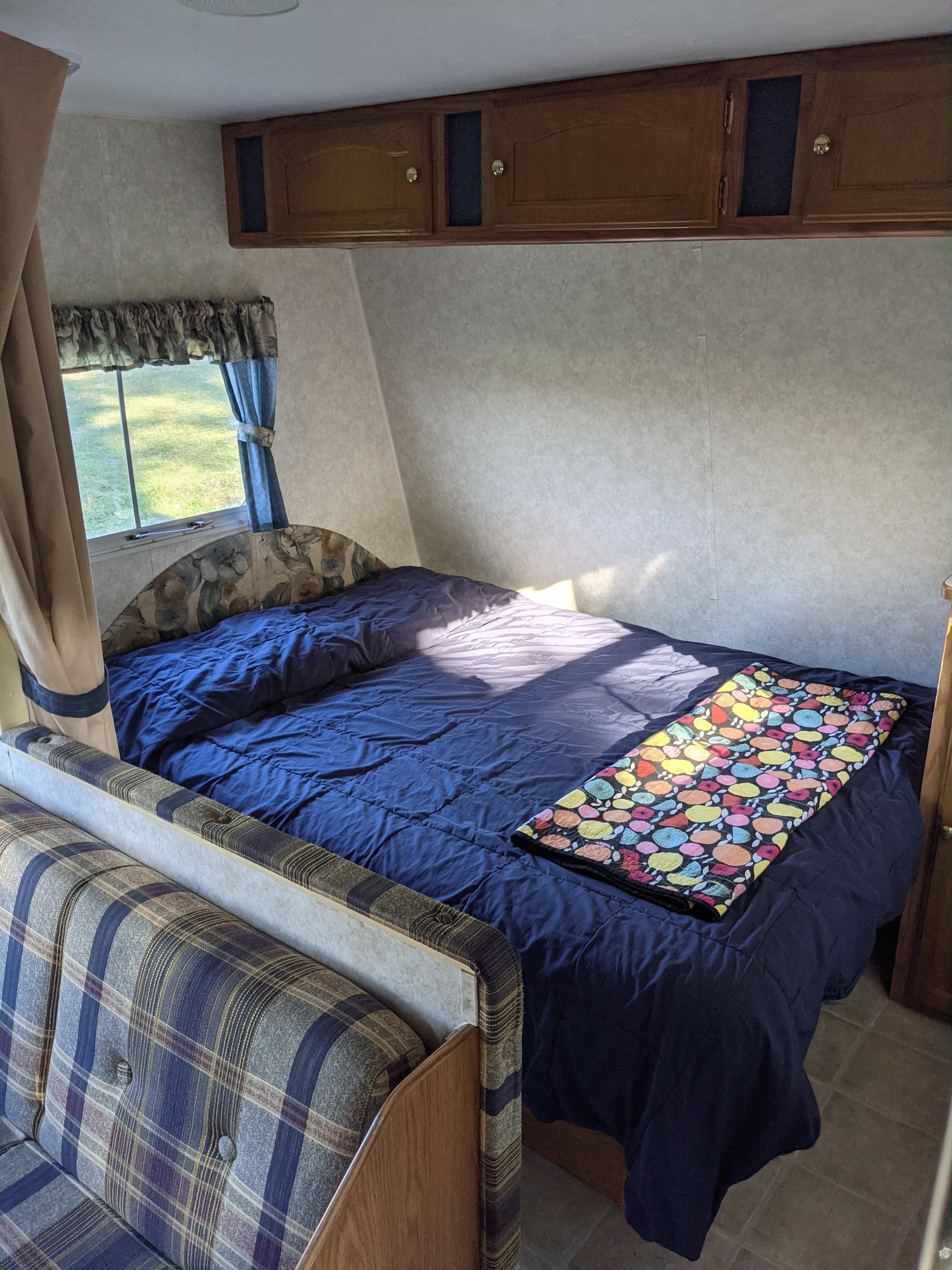 RV rental bedroom: cozy bed with blue comforter, colorful blanket, wood cabinets, and window view.. Keystone RV Springdale 2003