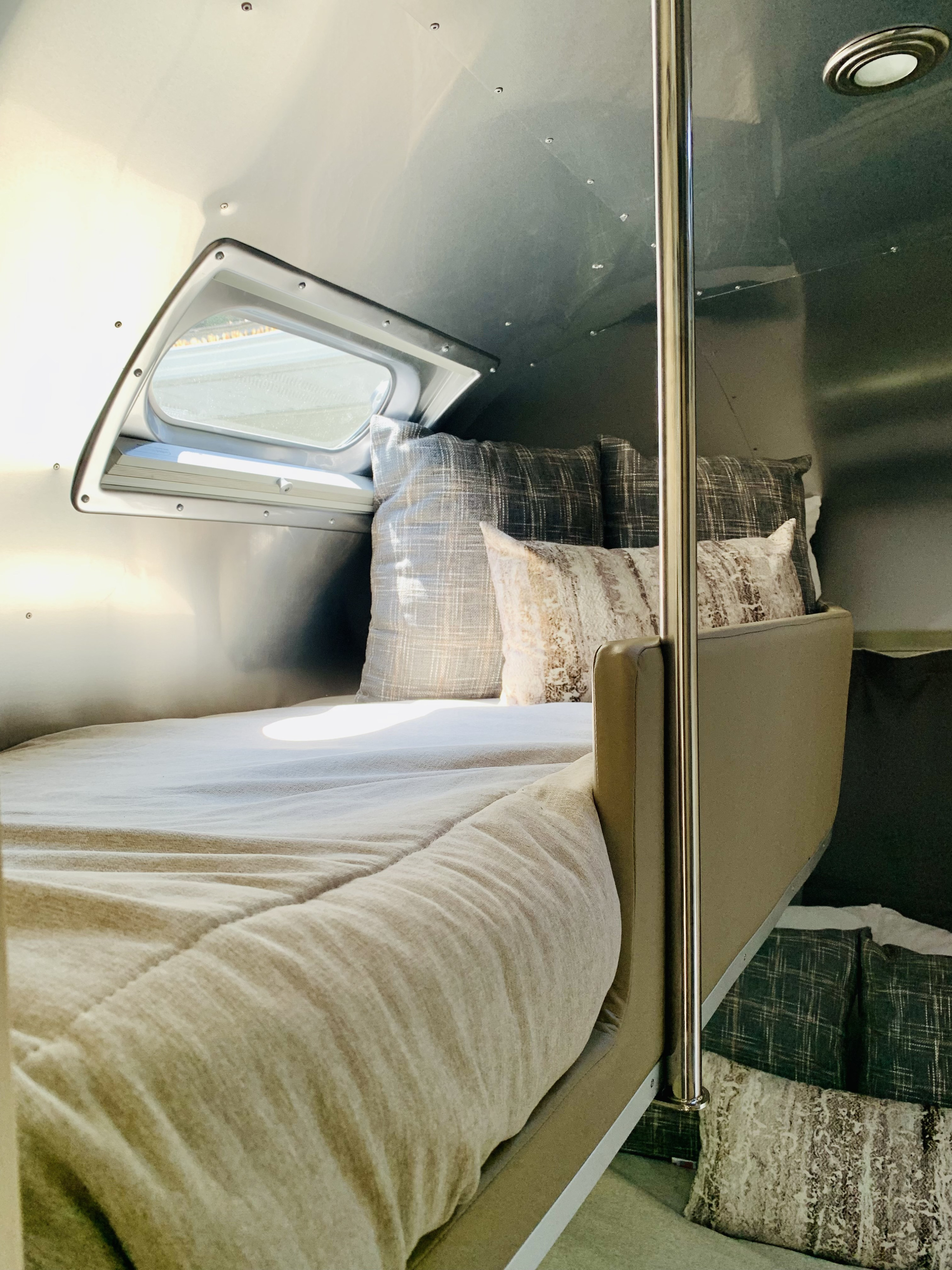 Inviting RV rental interior: a plush bed with patterned pillows, bright window, and sleek metallic walls. A second bunk is visible.. Airstream Flying Cloud 2022