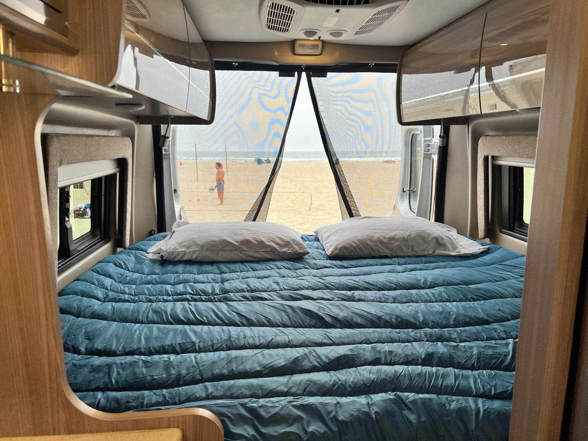 Inside a comfy RV rental, featuring a bed with a direct beach and ocean view through the open rear.. Thor Motor Coach Sequence 2022