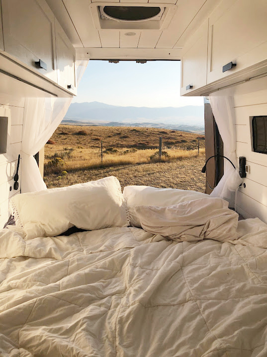 Cozy RV rental bed overlooks rolling hills and distant mountains, offering a serene mobile escape.. Mercedes-Benz Sprinter 2015