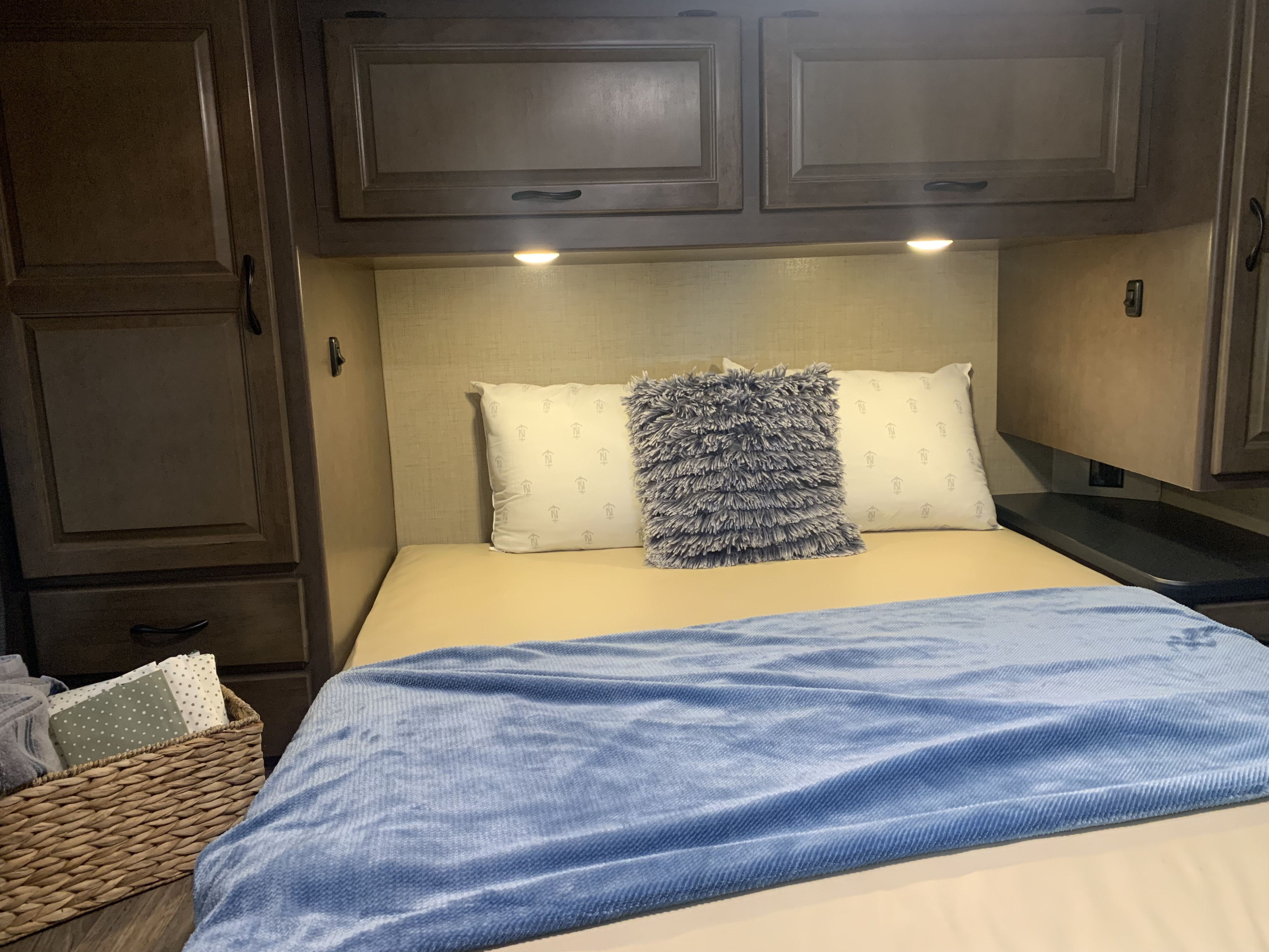 Cozy RV rental bedroom with a neatly made bed, soft blue blanket, pillows, warm lighting, and overhead storage cabinets.. Thor Majestic 2018