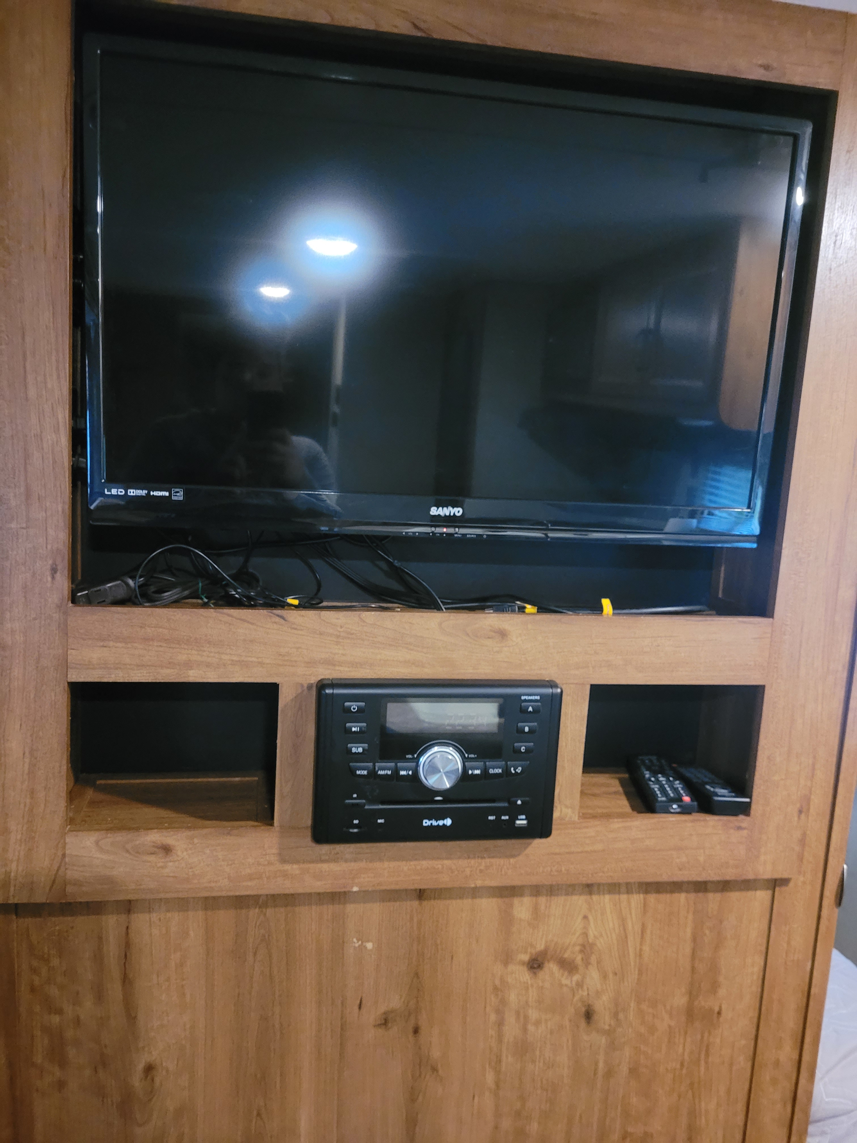RV rental entertainment system: a black Sanyo TV, Drive stereo, and remotes housed in a wood-paneled cabinet.. Gulf Stream Ameri-Lite 2017