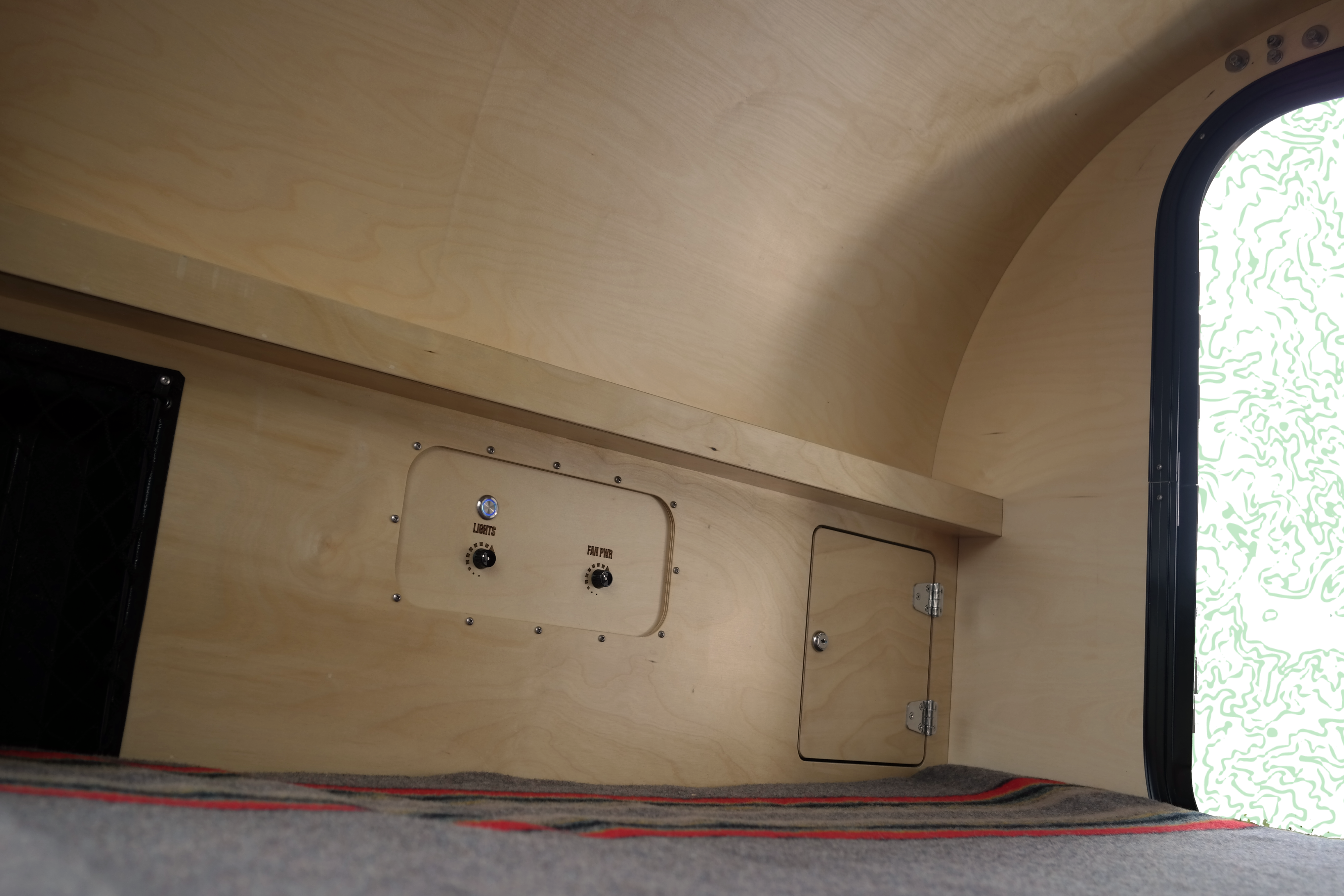 Inside an RV rental: Blonde wood interior with 'LIGHTS' & 'FAN PWR' controls, a small storage door, and cozy bedding.. Oregon Trail'R DoDrop ALPHA 2021