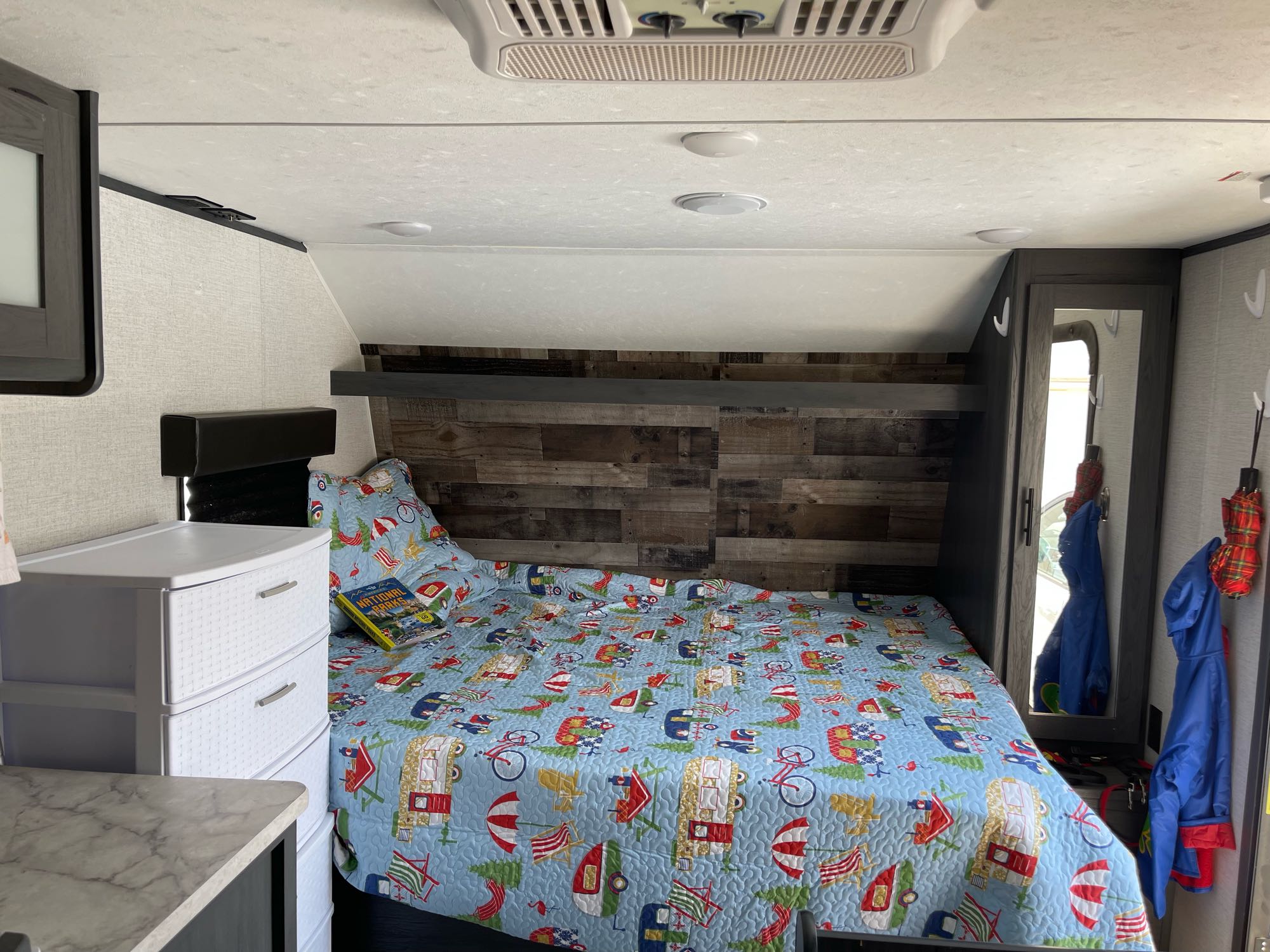 Cozy RV rental bedroom featuring a colorful camping-themed quilt, white storage drawers, and a mirror.. Dutchmen Aspen Trailer 2021