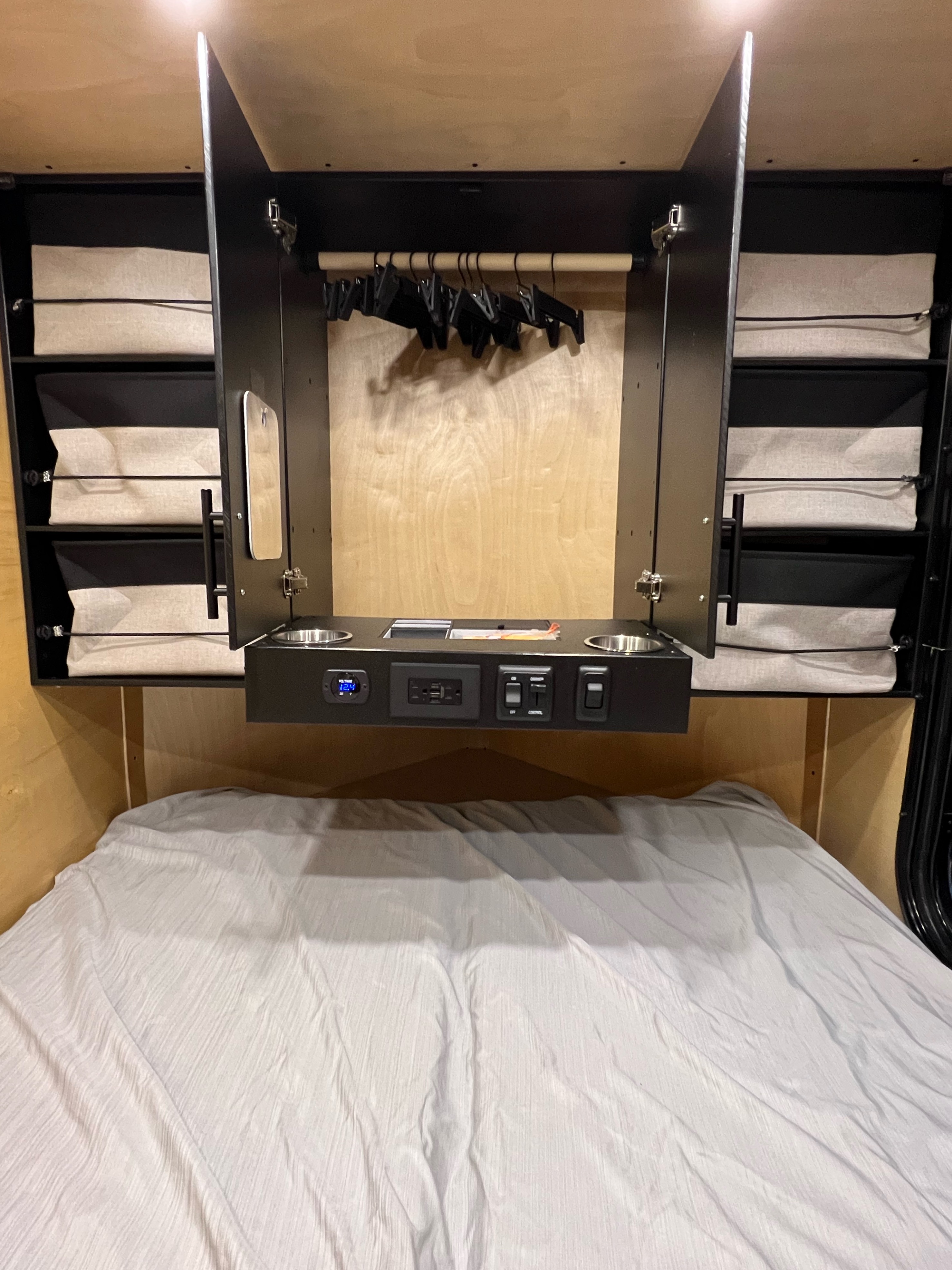 An organized RV rental interior showing a bed with white sheets, overhead storage with hangers, cup holders, and power controls.. Custom Travel Trailer 2022