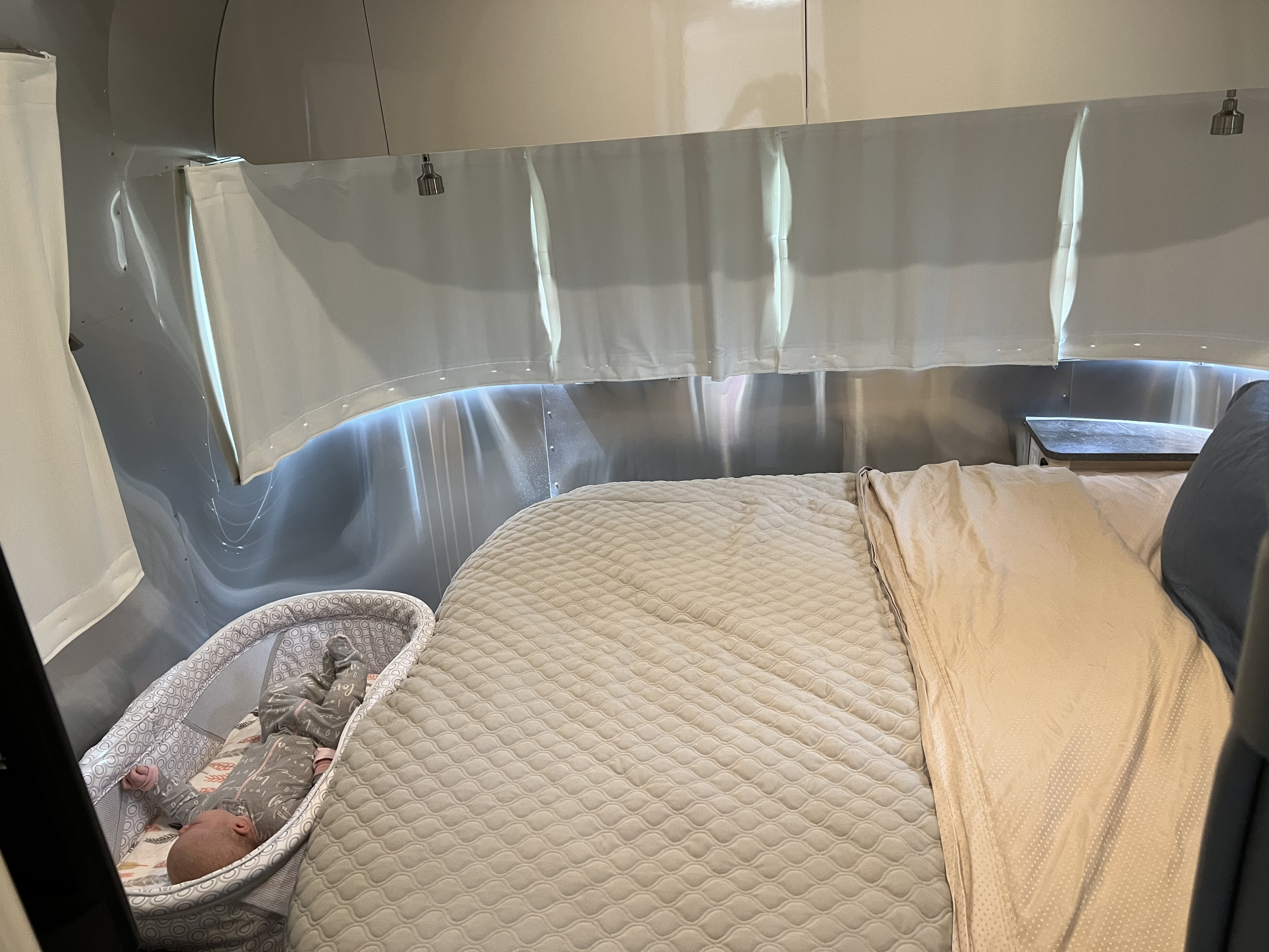 Inside an RV rental: a bright bedroom with quilted bed, baby in bassinet, and aluminum walls. Ideal for family travel.. Airstream Flying Cloud 25RB 2022