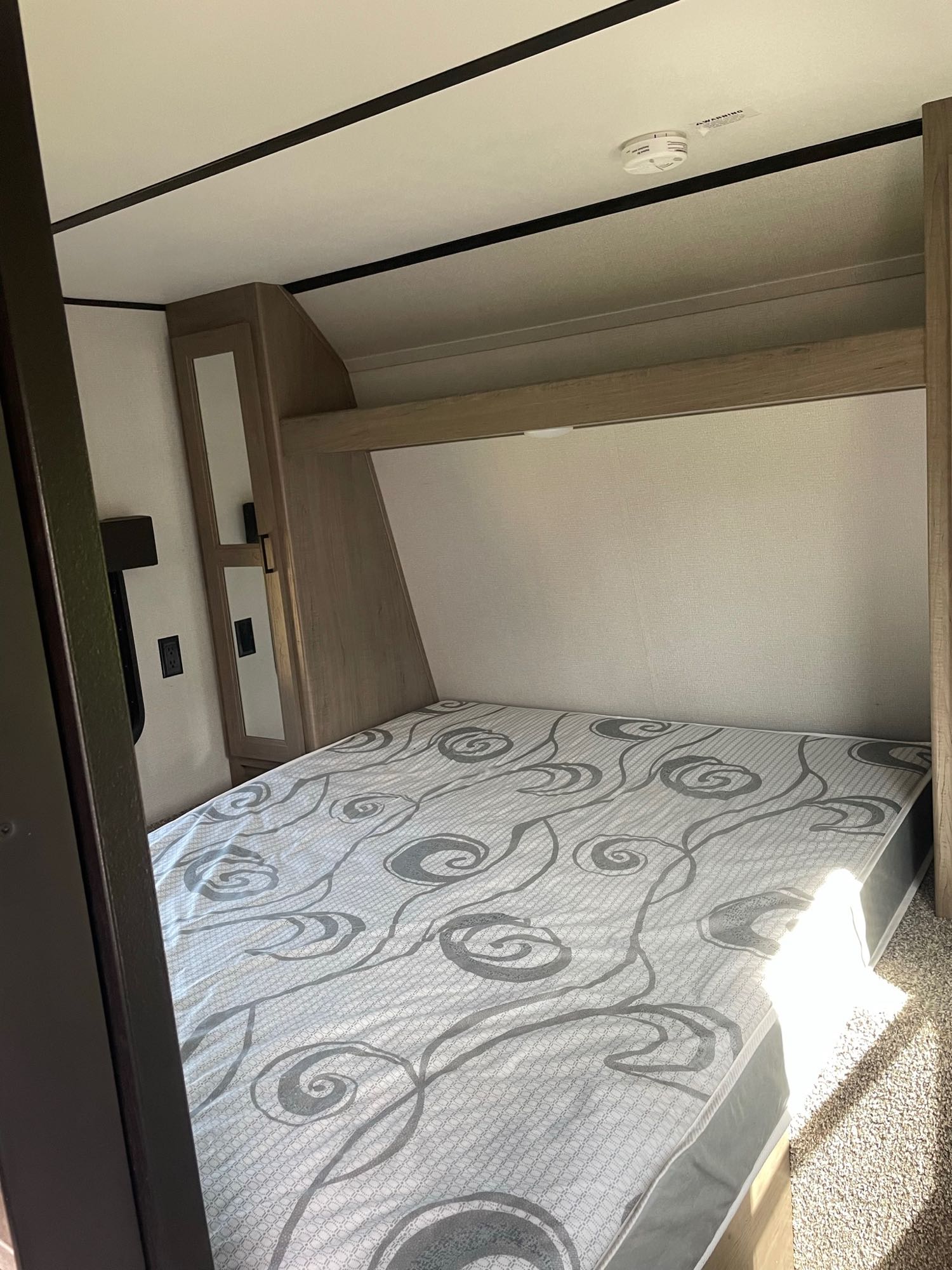 Inside an RV rental, a bedroom with a patterned mattress, light wood cabinetry, and a ceiling smoke detector.. Keystone RV Hideout 2020
