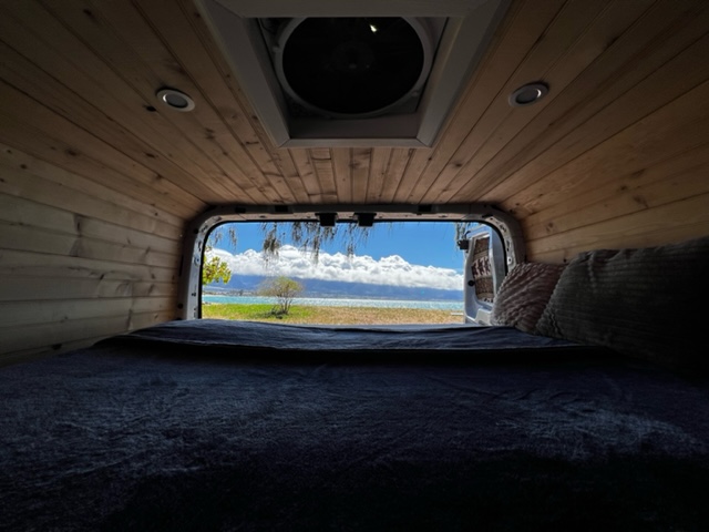 Cozy RV rental interior with bed & wood paneling. Stunning view of turquoise water, blue sky, & coastal trees. Perfect travel!. Custom Ford Transit Van 250 2015