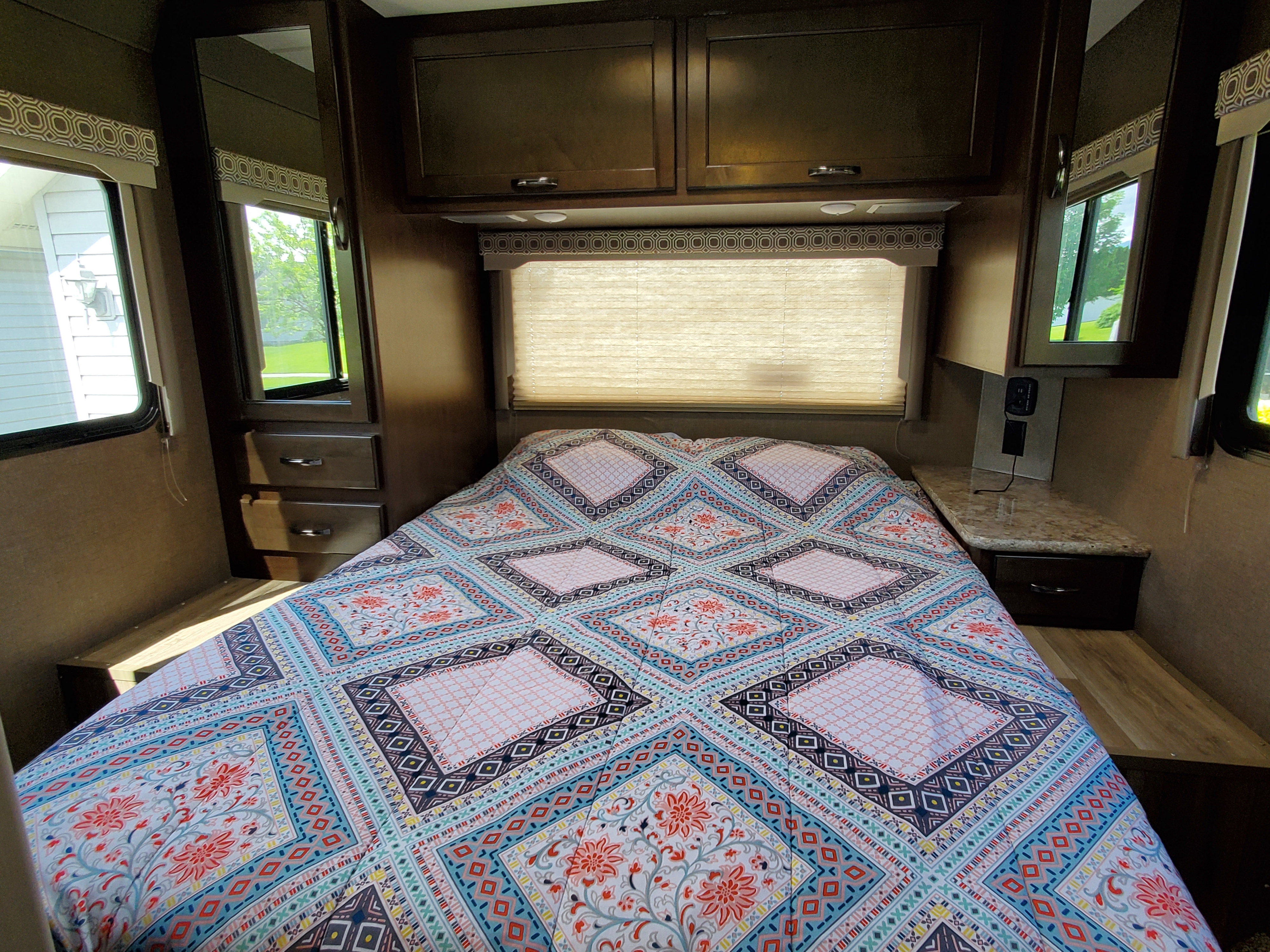 A colorful patterned quilt covers the bed in this inviting RV rental bedroom, flanked by dark cabinets & sunny windows.. Thor Four Winds 2018