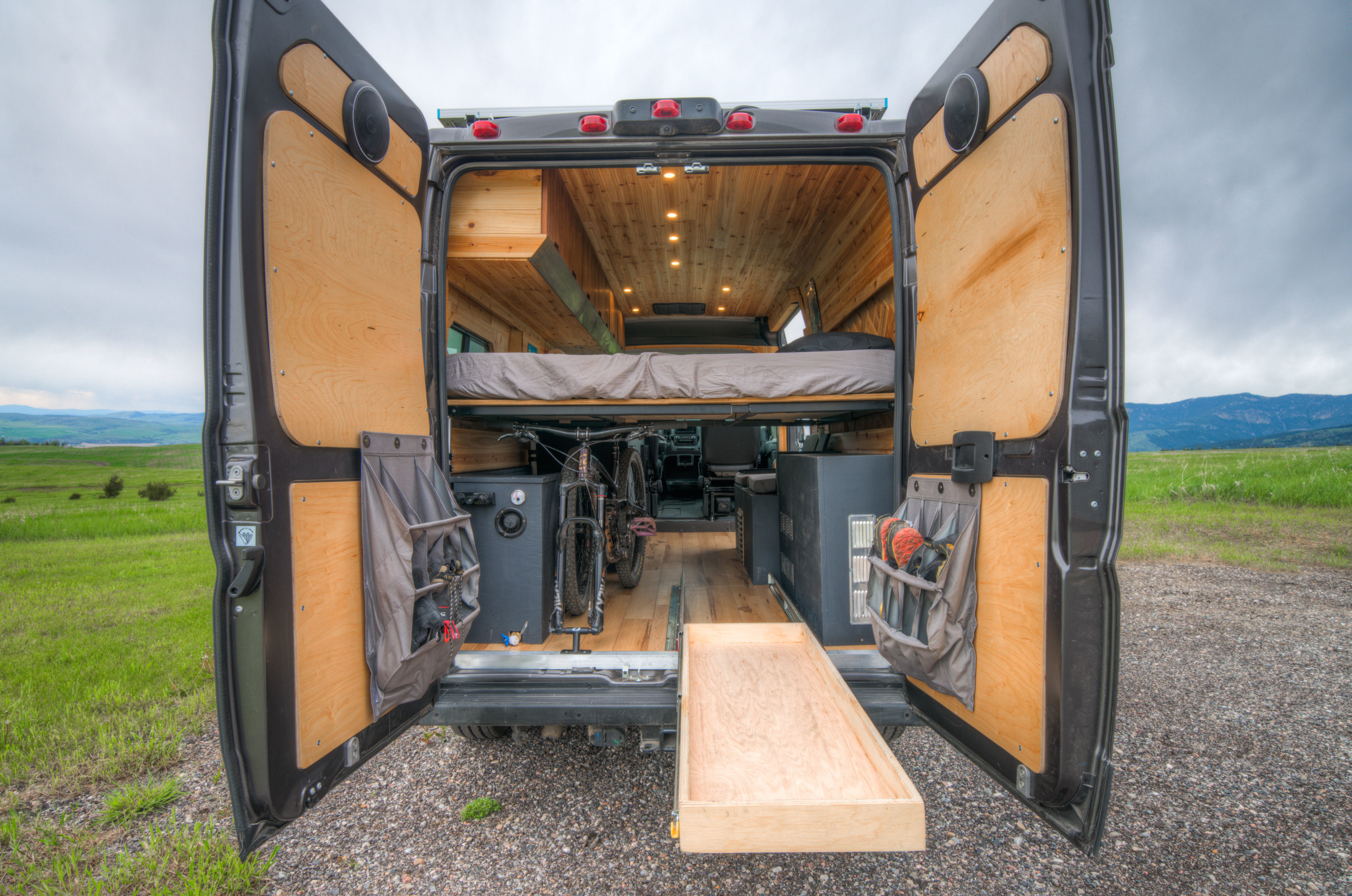 RV rental: Rear view of a custom camper van, doors open, revealing a wood interior, bed, bike storage & pull-out tray in nature.. Ram Promaster 2022
