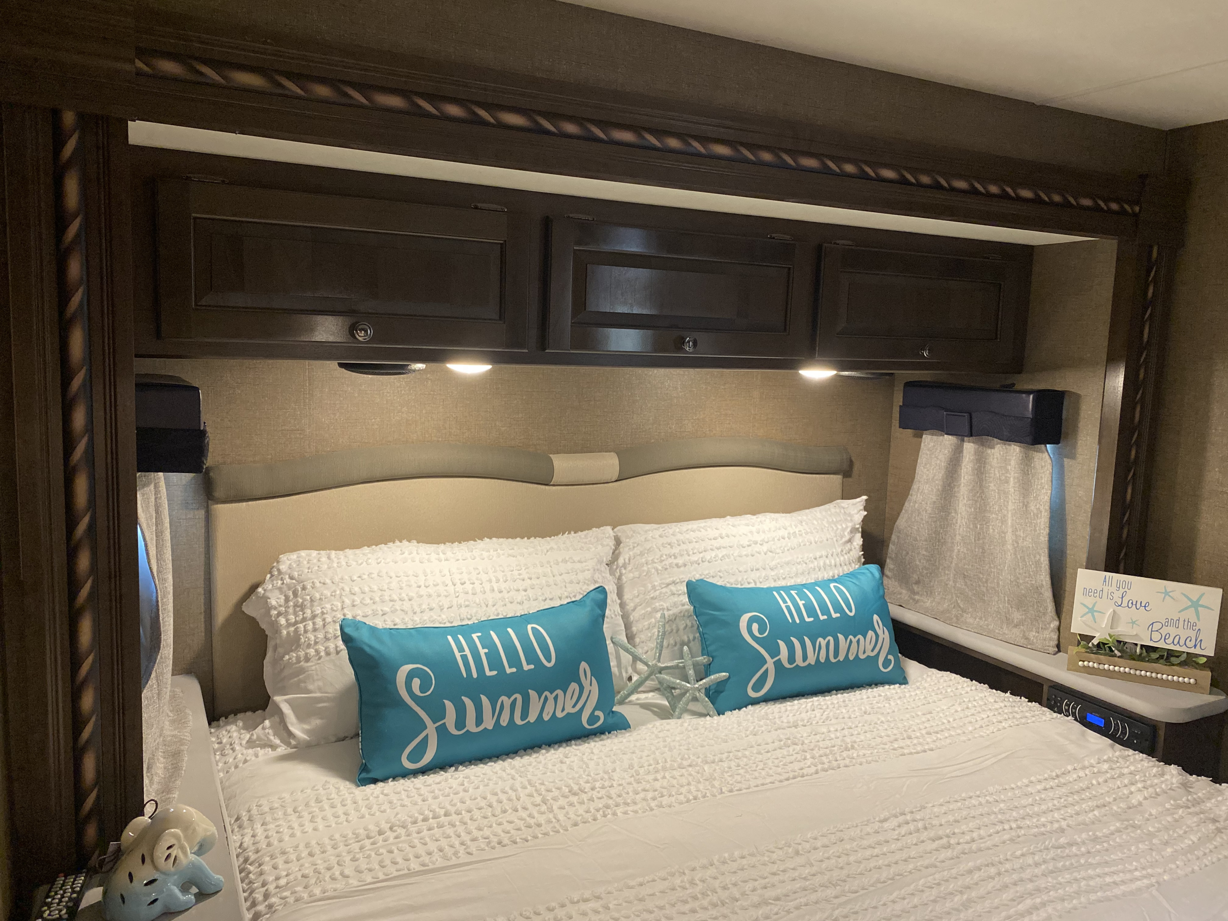 Cozy RV rental bedroom with white bedding, blue "HELLO Summer" pillows, overhead cabinets, and beach decor for summer trips.. Thor Windsport 2017