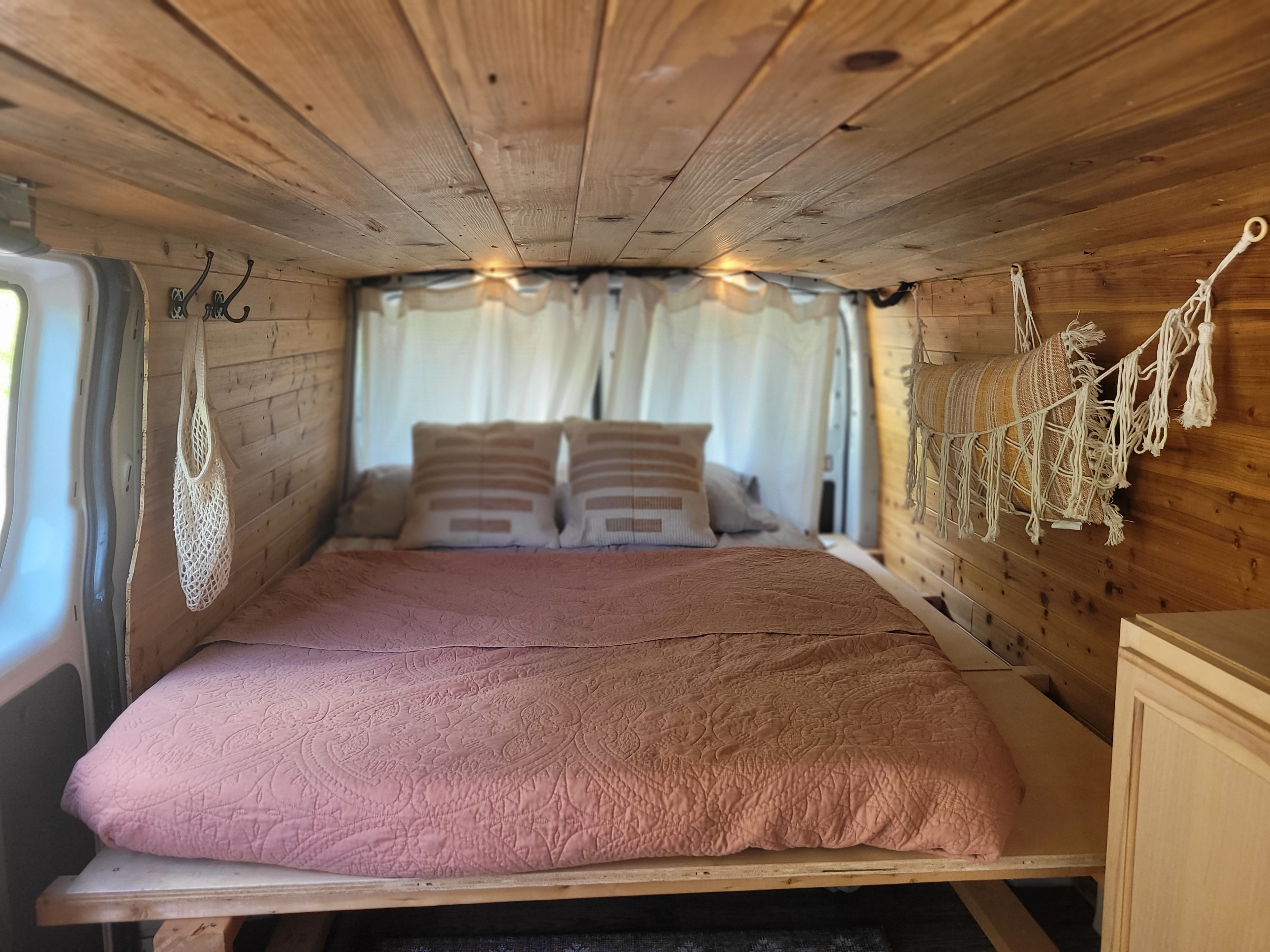 A cozy RV rental bedroom interior with light wood paneling, a pink bed, decorative pillows, and macrame accents.. Ford Transit 2016